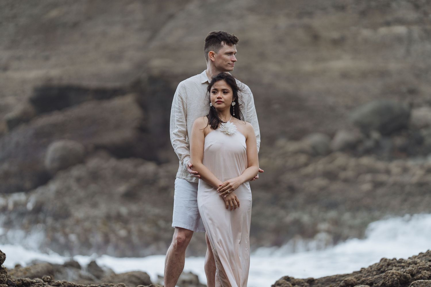 A romantic couple embraces against dramatic coastal cliffs and crashing waves.
