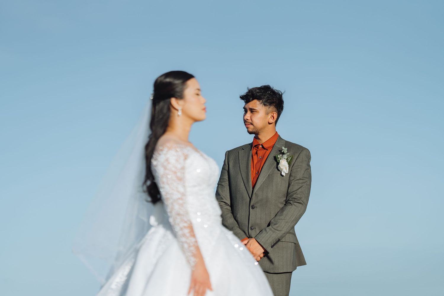A couple in wedding attire shares a tender moment against a bright blue sky.