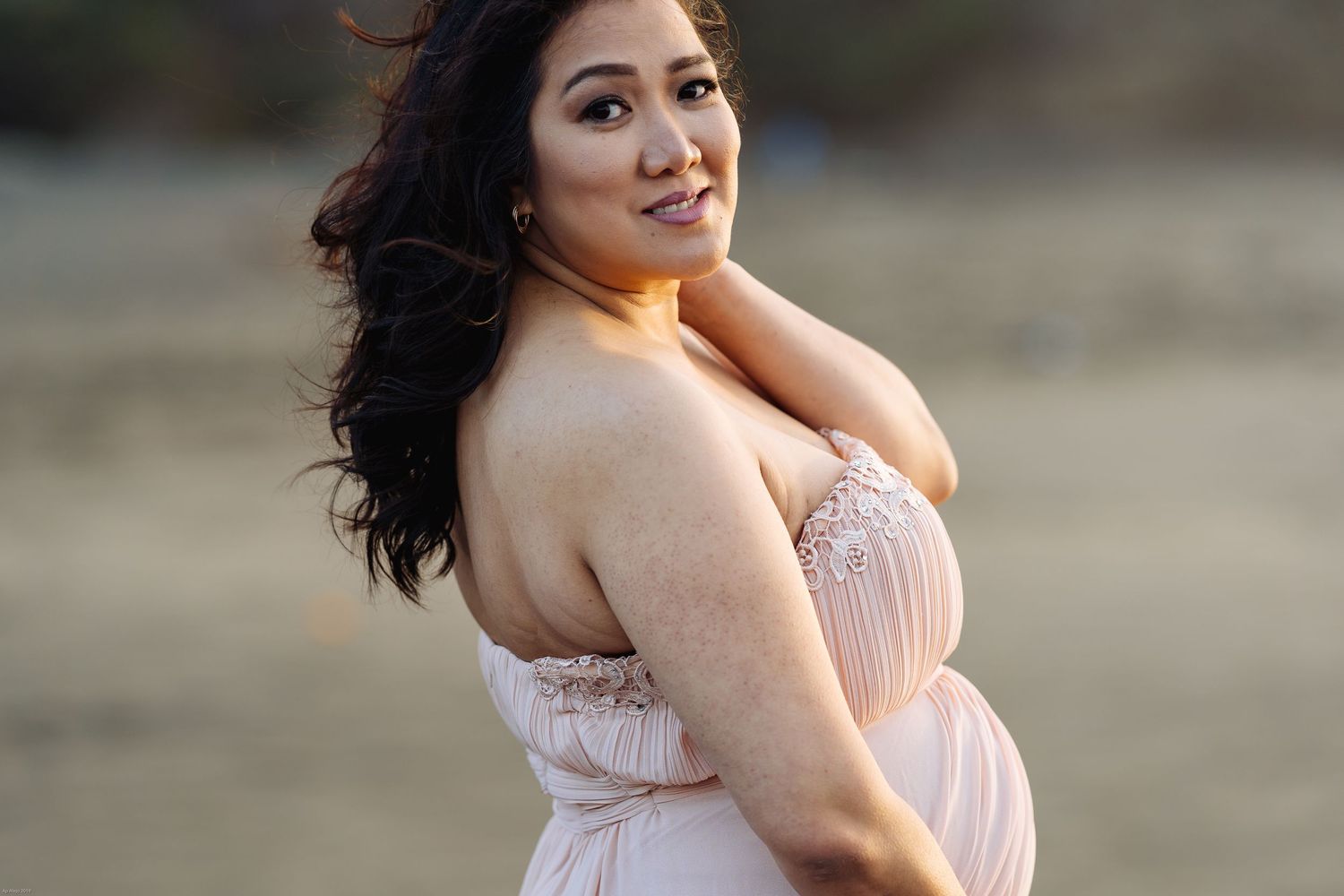 Side profile maternity portrait in a flowing pink dress against a natural backdrop.