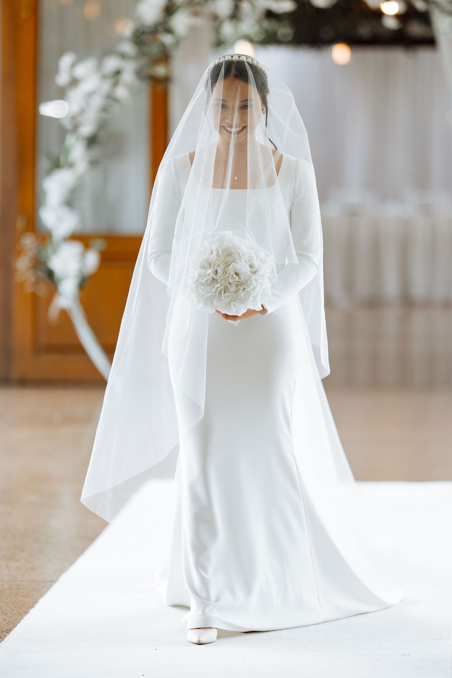 Elegant bride in minimalist white wedding gown and veil holds bouquet on white runway.