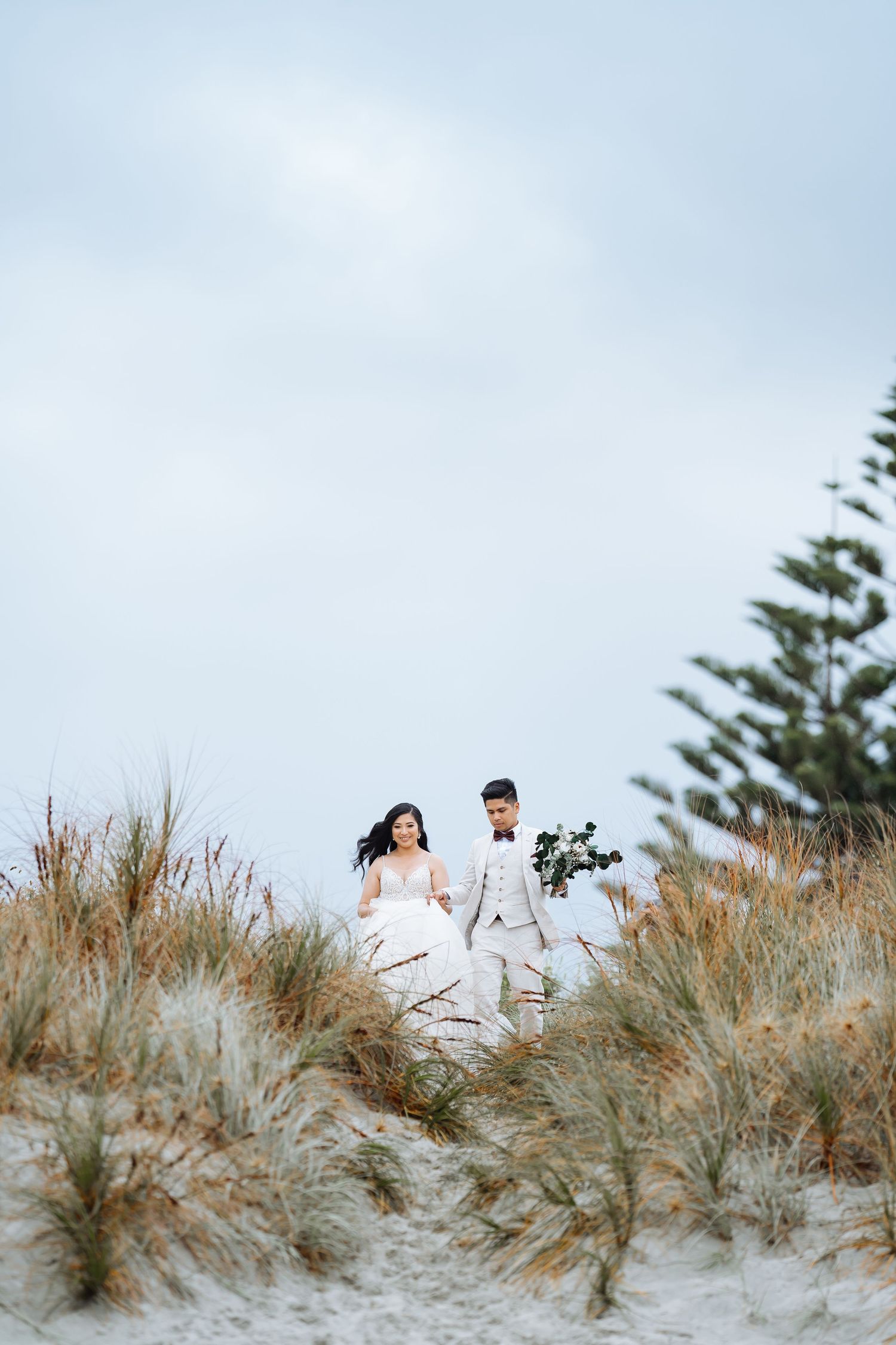 A couple walks through sandy dunes with pine trees in the distance.