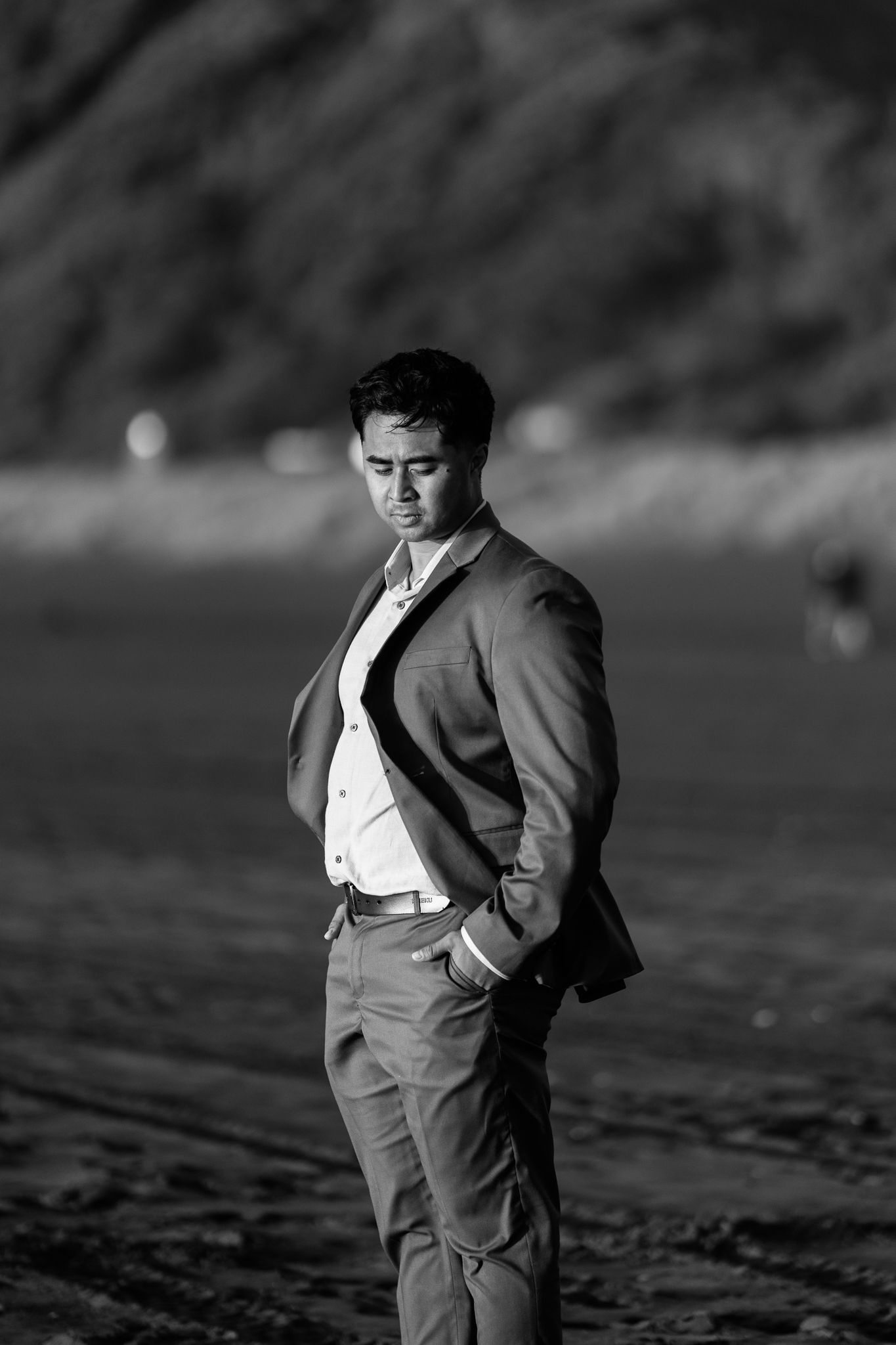 Black and white portrait of groom in suit on beach with dramatic lighting.