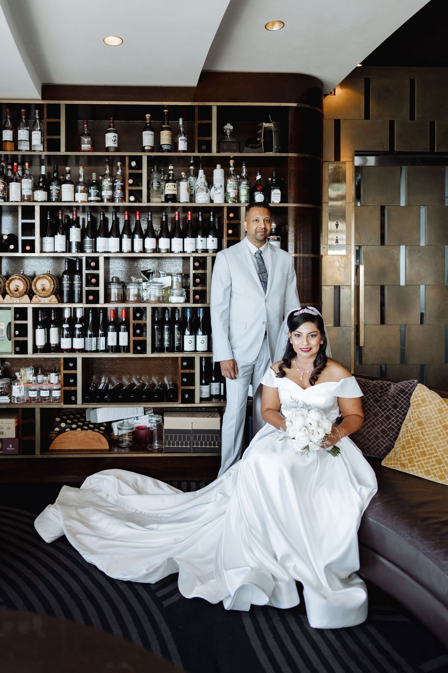Wedding couple poses in elegant bar lounge with extensive liquor display shelving and modern decor.