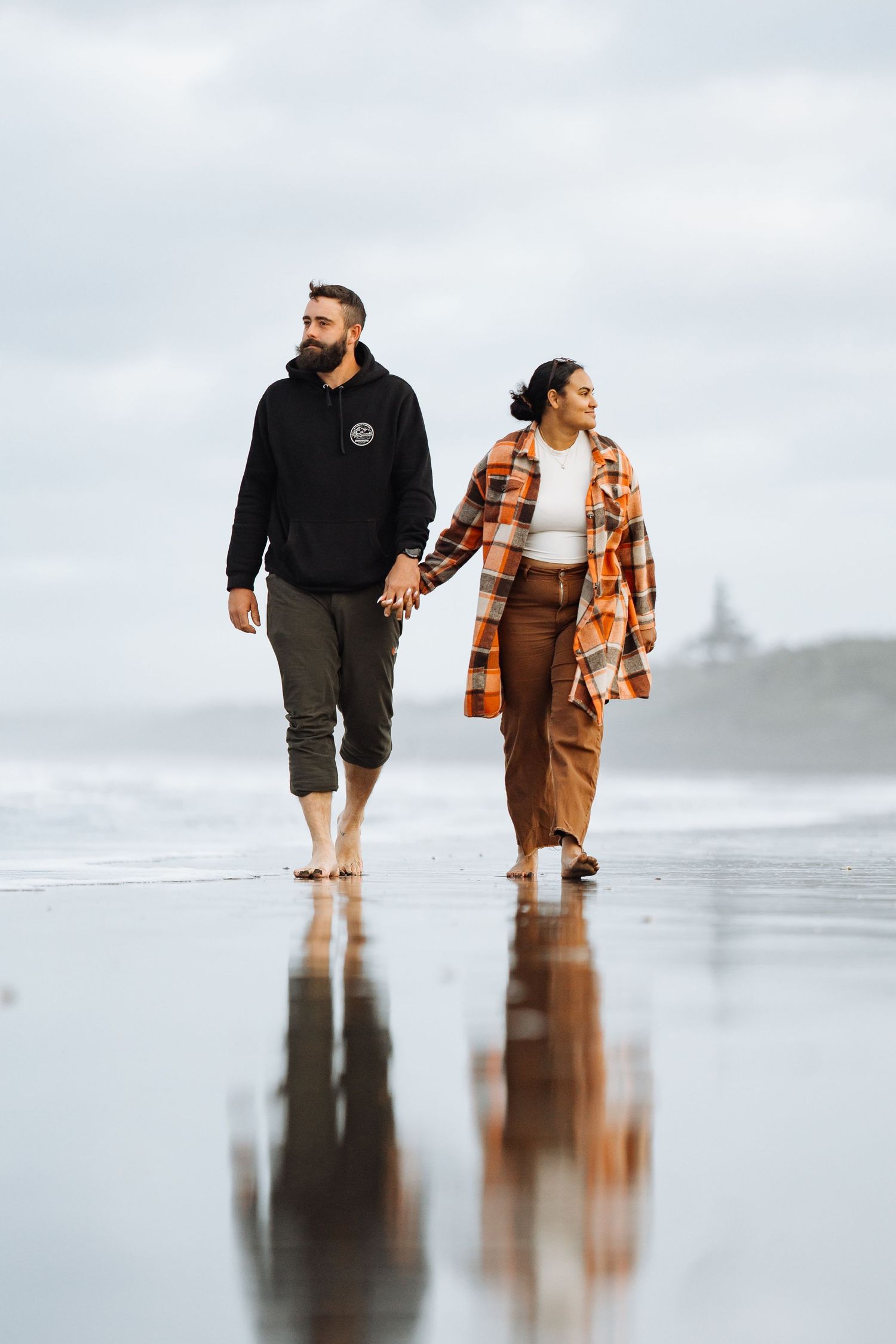 Couple walking hand-in-hand barefoot on a reflective black sand beach, man in black hoodie, woman in plaid jacket.