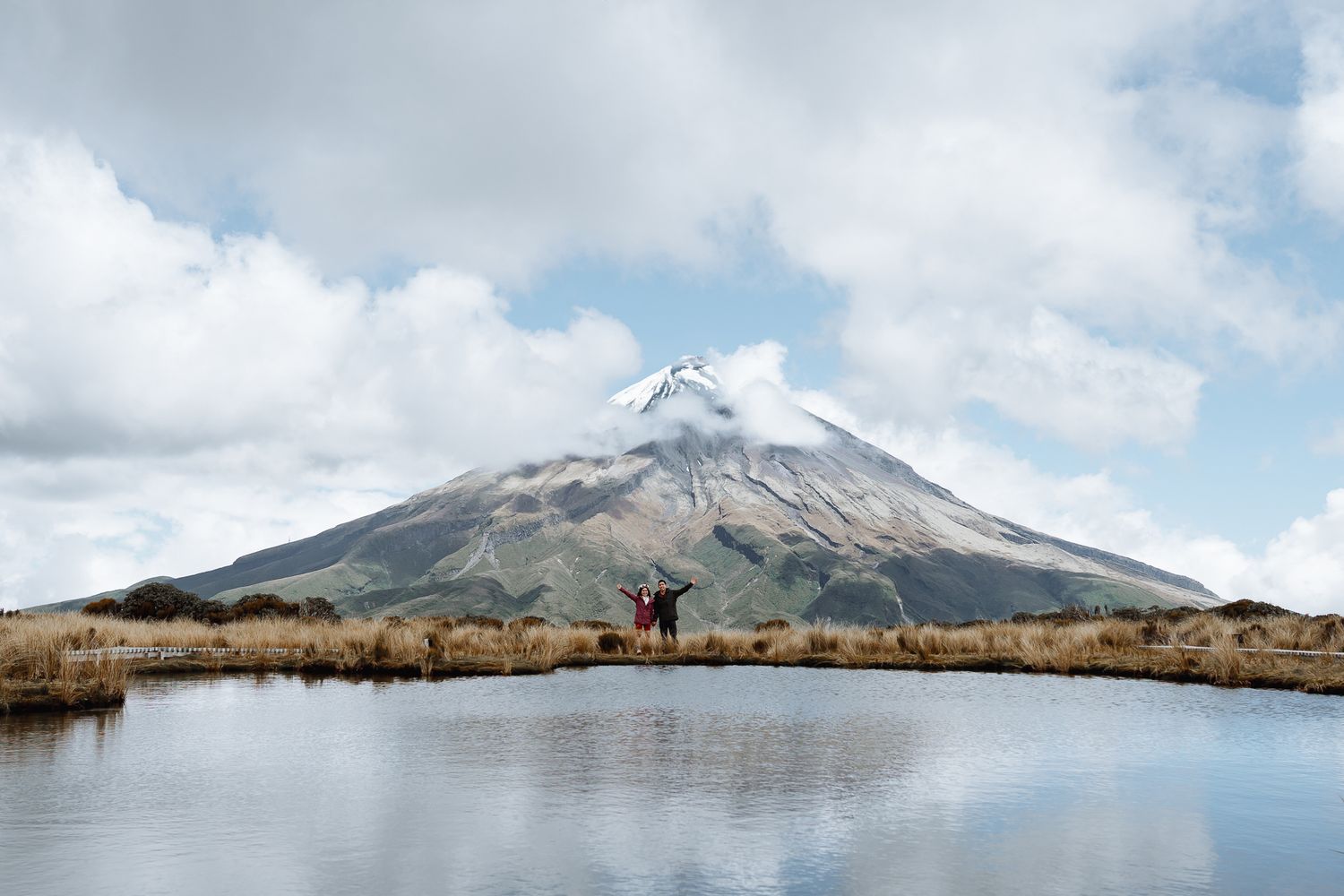 Snow-capped mountain peak reflects in still lake waters under cloudy sky.