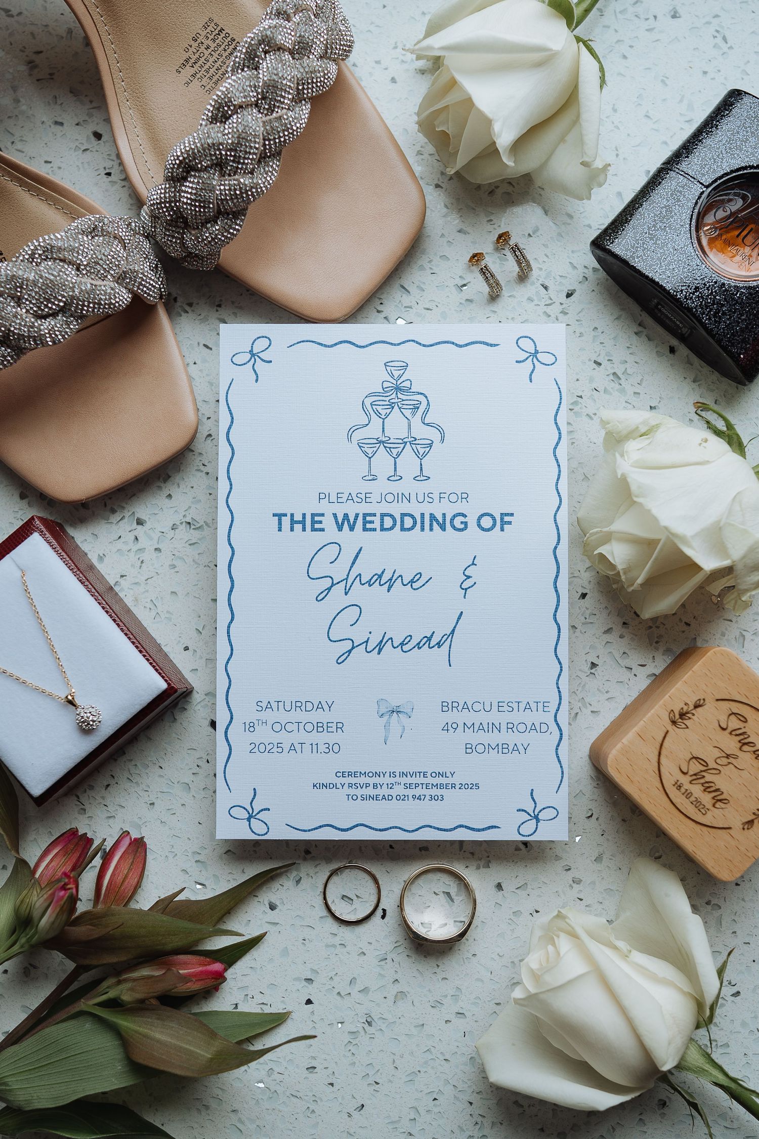 Wedding invitation flatlay with braided sandals, white roses, and wedding rings on textured surface.