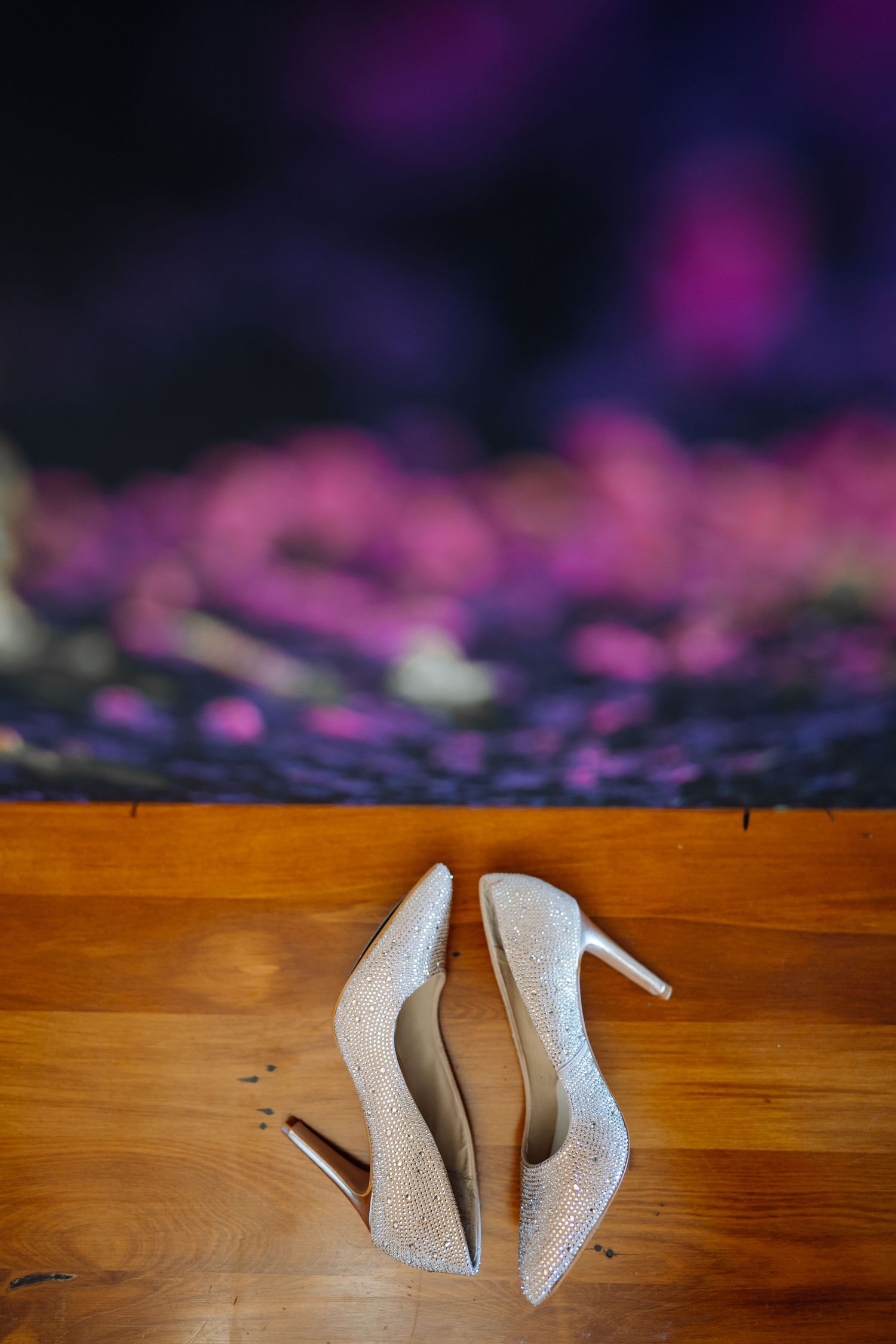 Sparkly white wedding heels photographed on wooden flooring with purple bokeh lighting effects.