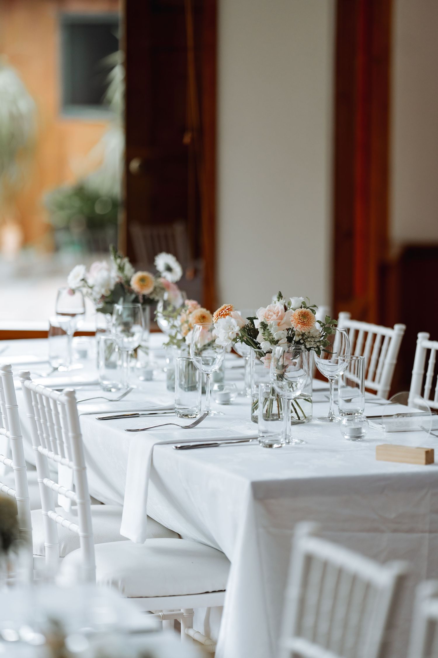 White and blush floral centerpieces adorn tables at an elegant wedding reception with white chairs.