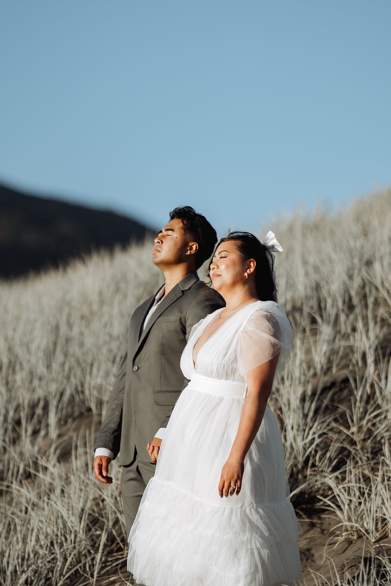 A couple in wedding attire stands in tall beach grass against a bright blue sky.
