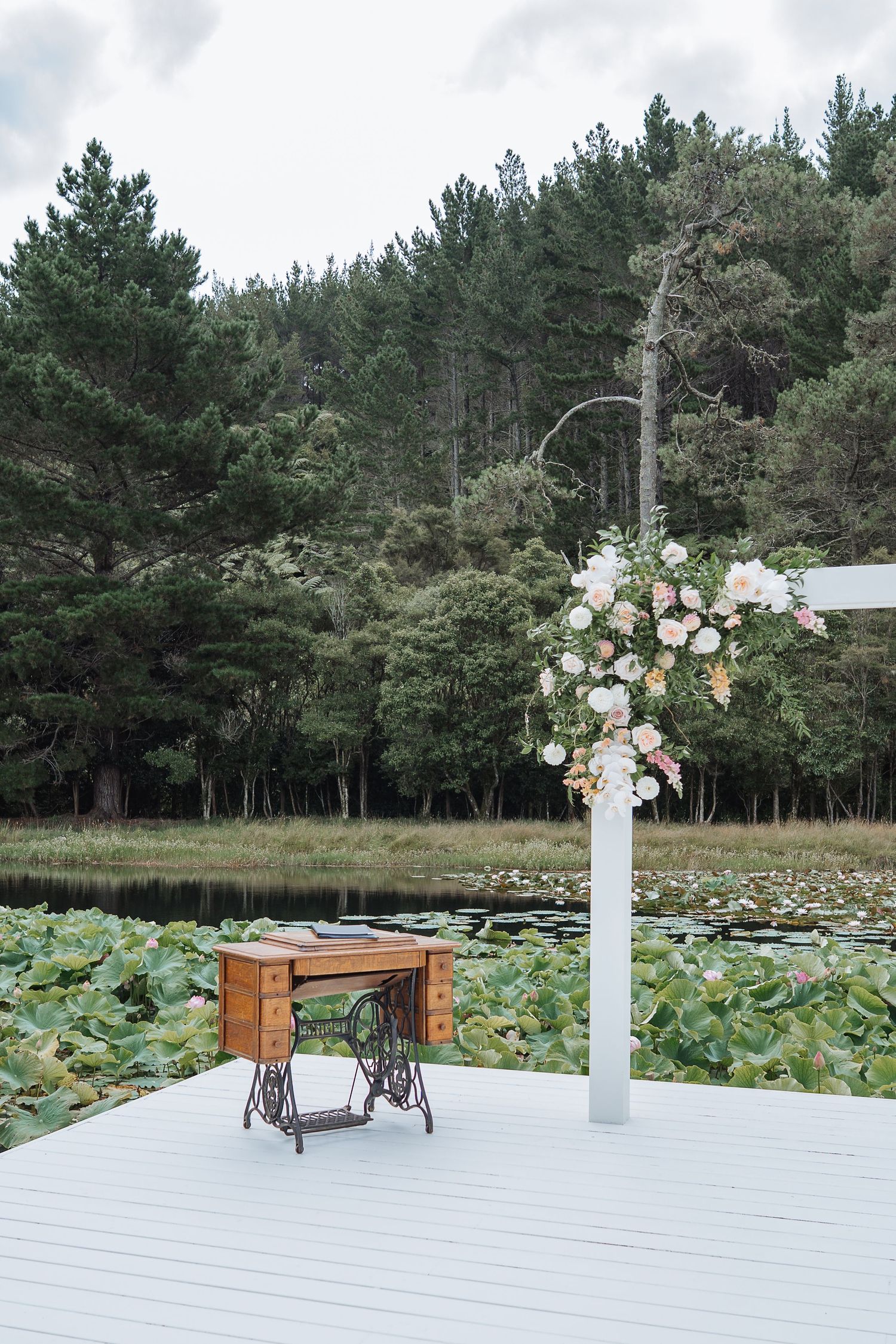 Outdoor wedding ceremony setup with white deck, floral arrangements and pine trees in background.