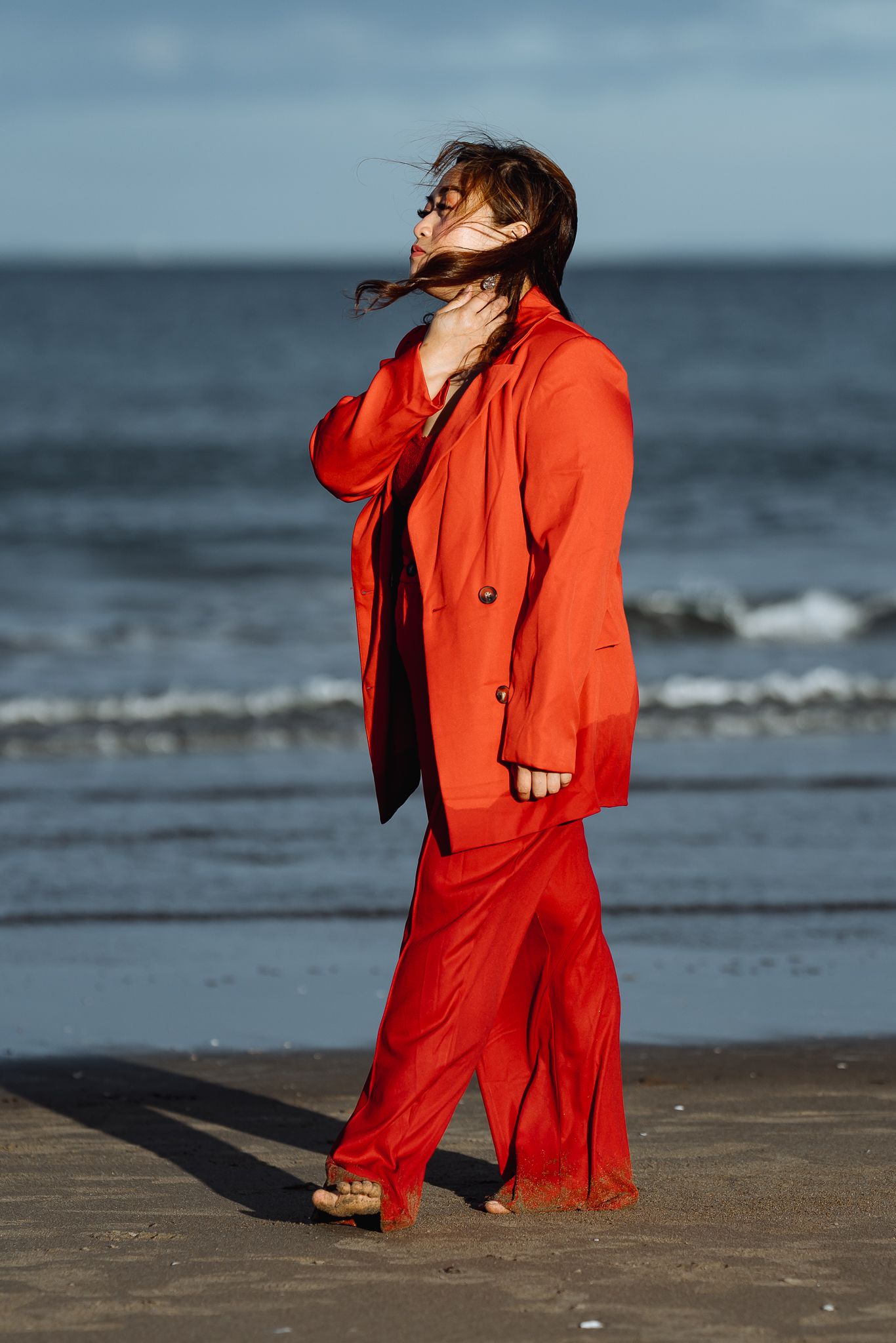 A figure in a vibrant red suit poses dramatically on a beach with windswept hair against an ocean backdrop.