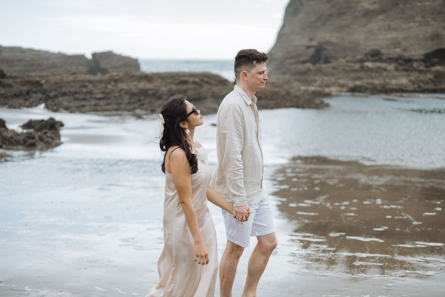 Couple in light colored outfits standing together on rocky beach coastline.