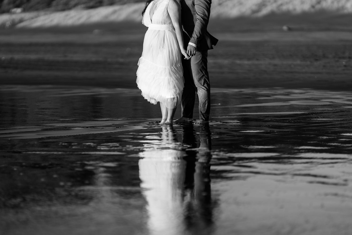 A black and white reflection of a couple walking in shallow beach water.