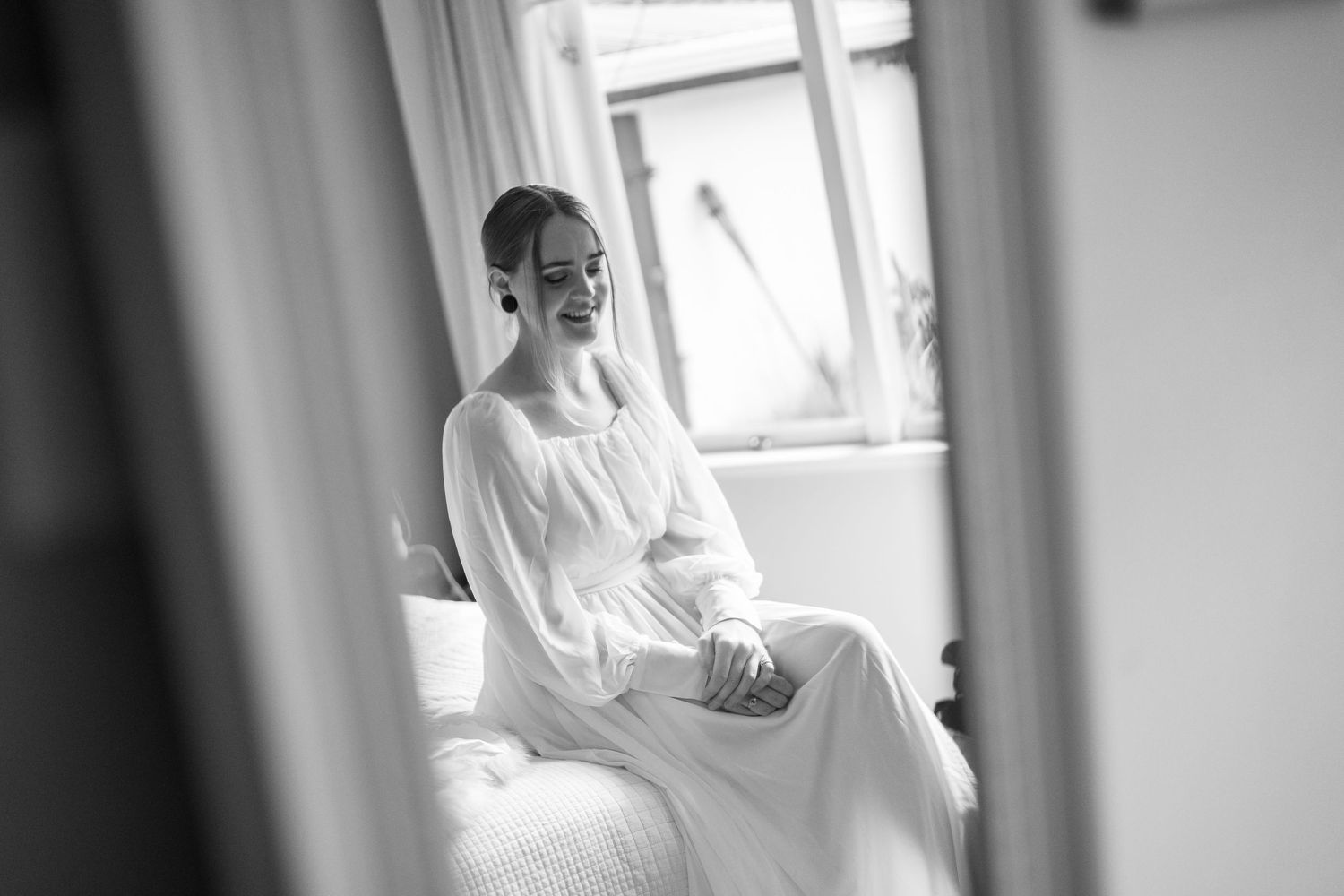 A black and white portrait of a bride sitting by a window in quiet contemplation.