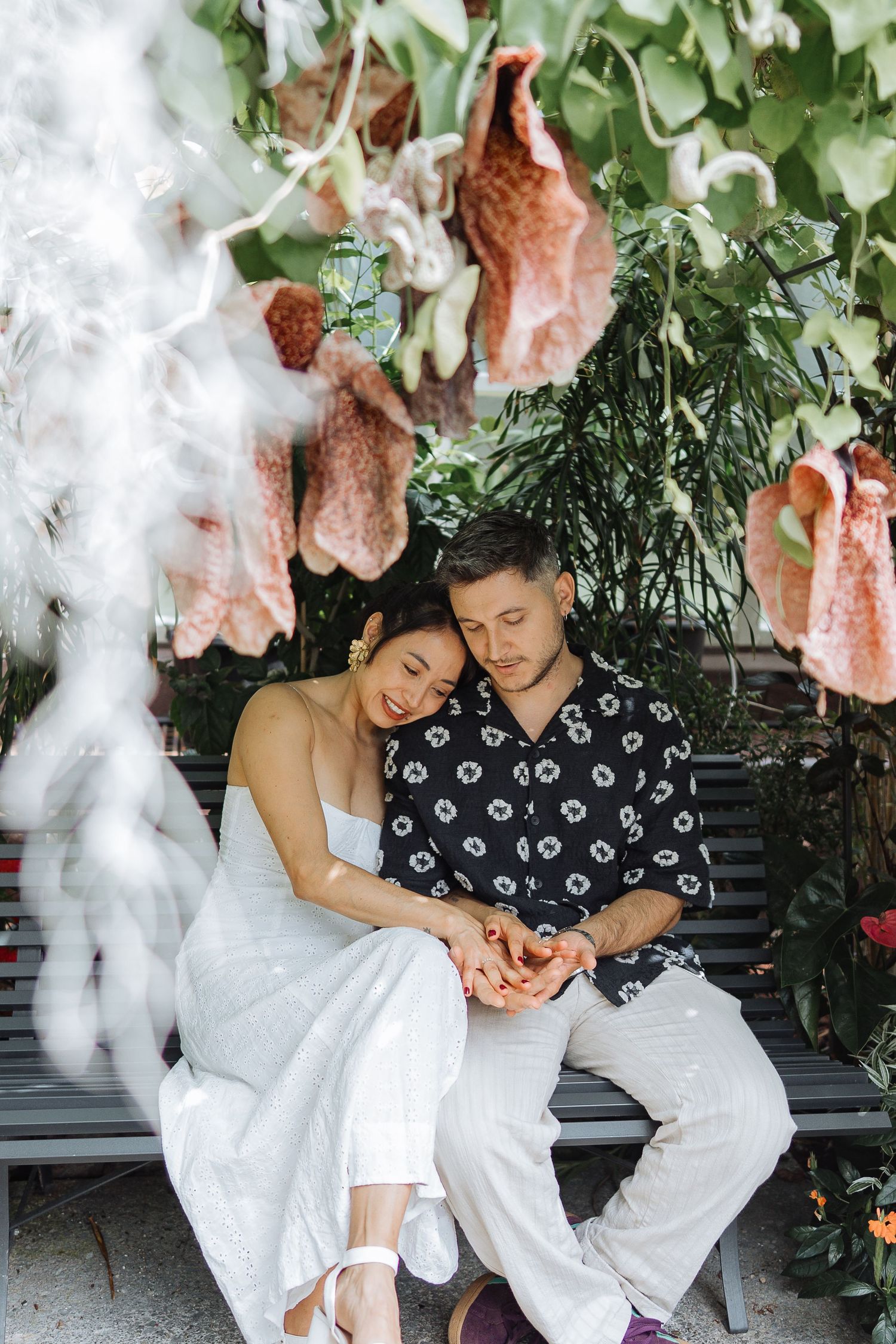 A couple shares a tender moment on a bench surrounded by pink and white flowers.