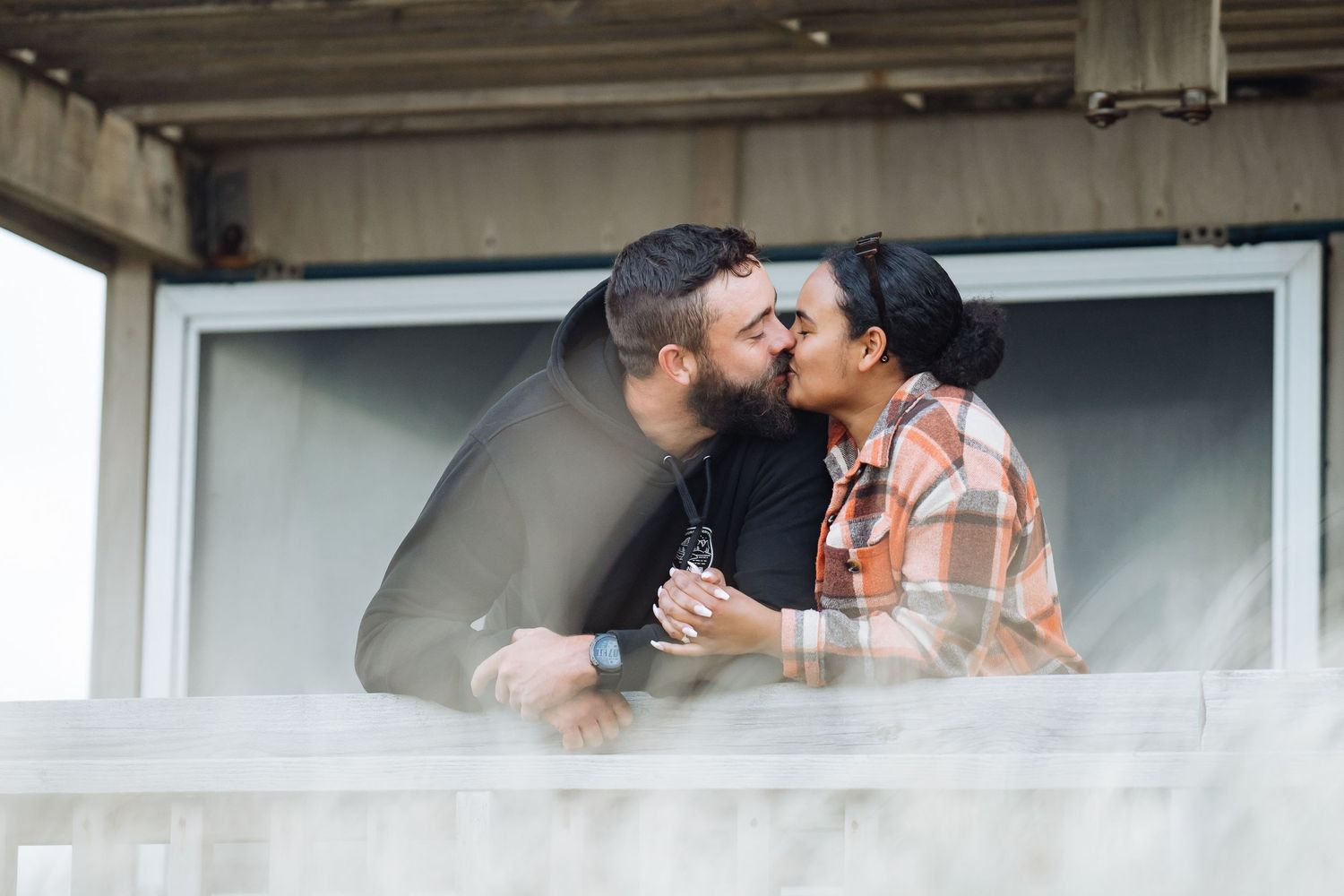 Couple sharing a romantic kiss on a balcony, man in dark jacket and woman in plaid shirt.