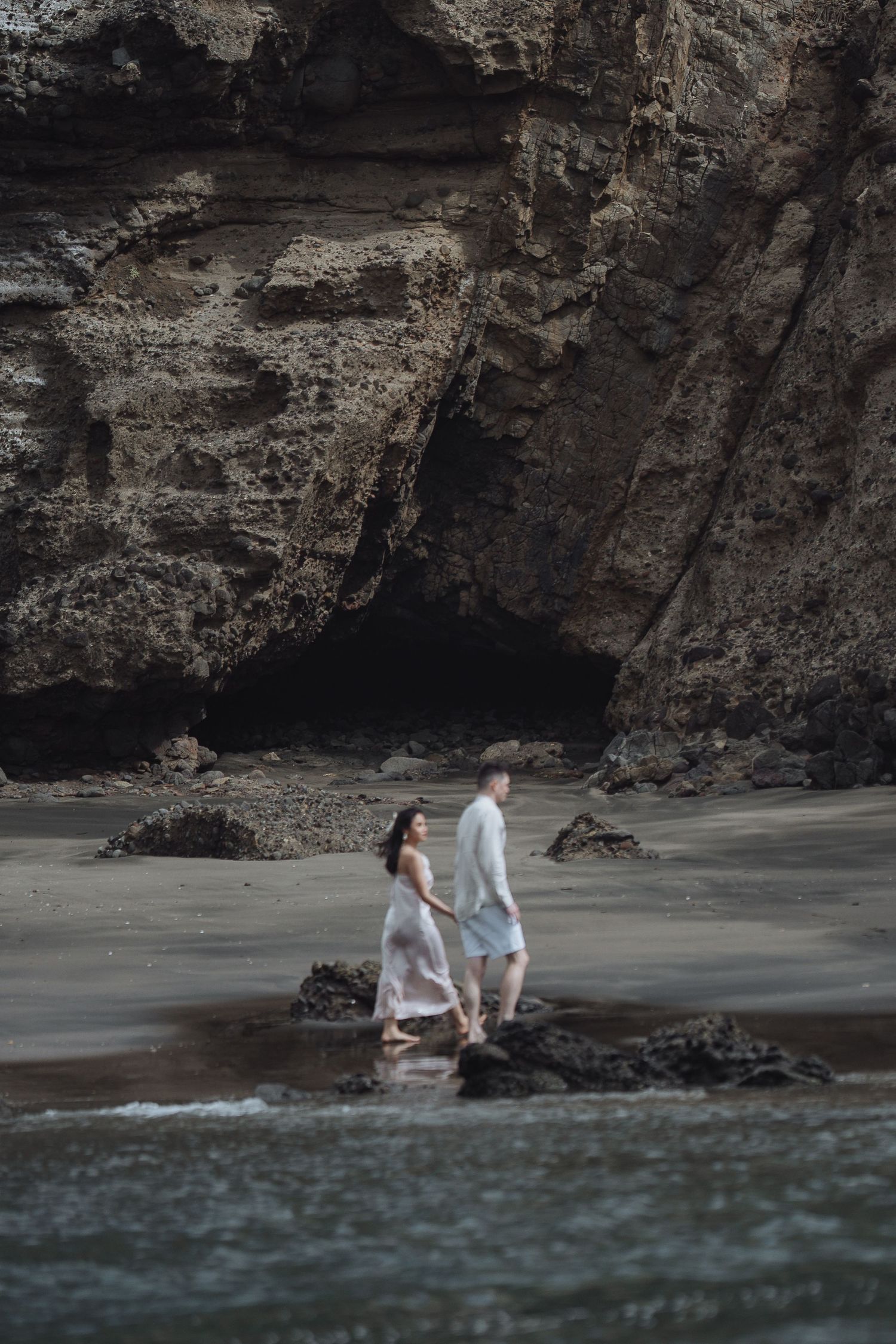 A couple walks hand in hand along a dramatic rocky beach with caves and cliffs.