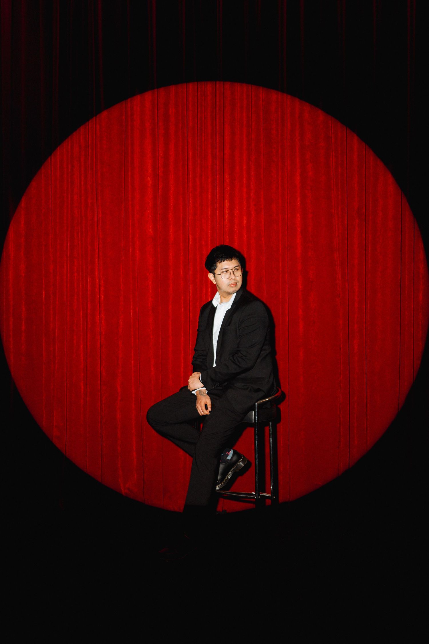 Person in black suit sits on stool against red curtain backdrop with spotlight.