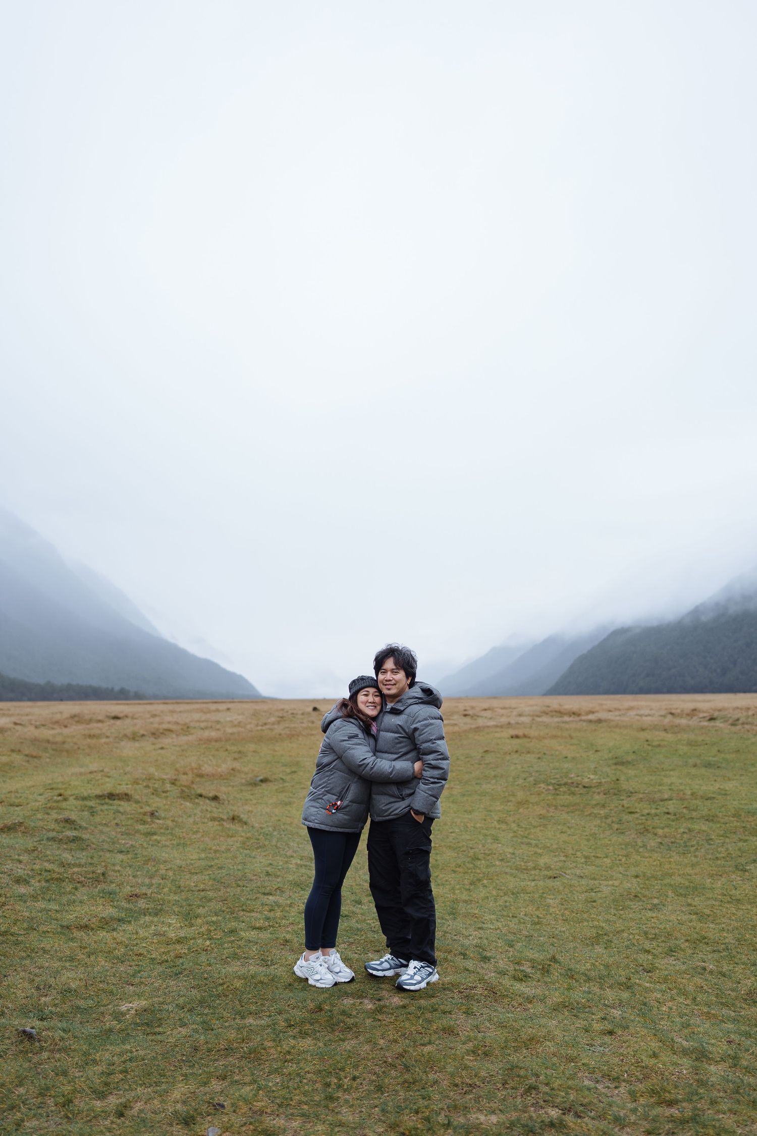 Two people in matching gray winter jackets embrace in a misty mountain valley.