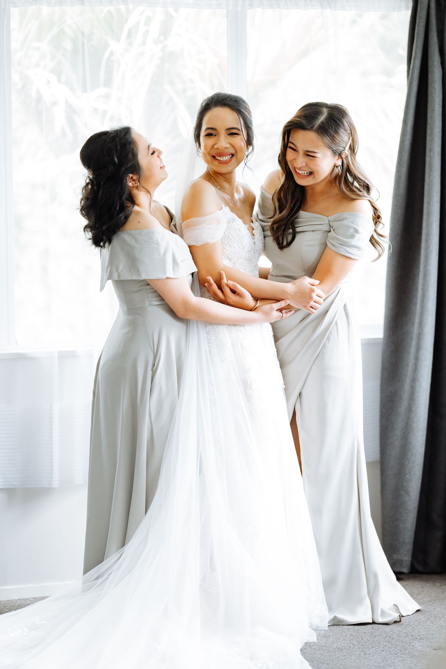 Three women in light-colored dresses share a joyful moment in a bright room.