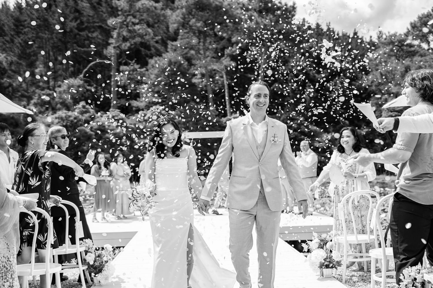 Joyful outdoor wedding celebration with guests throwing confetti in sunlight.