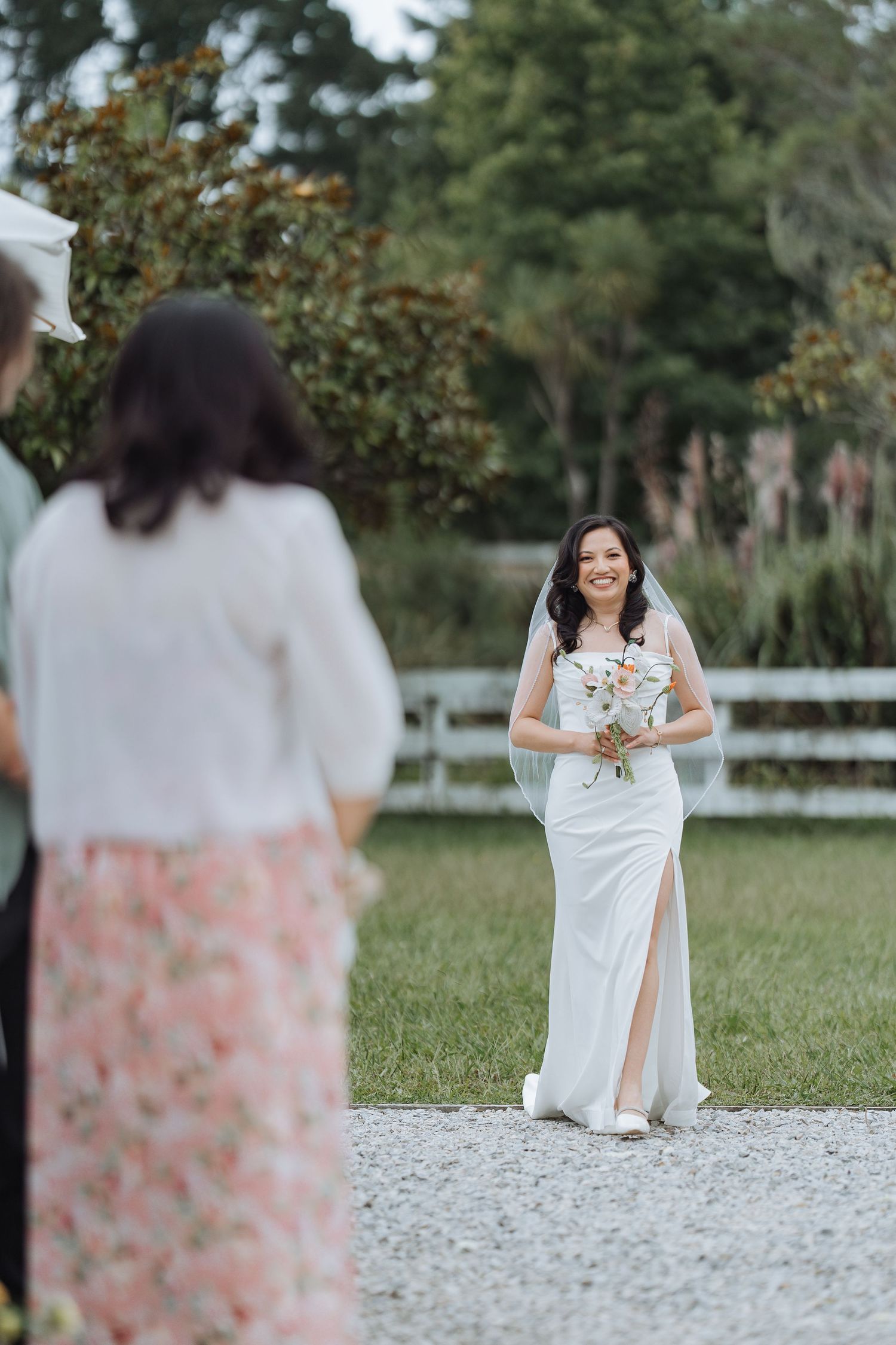 A bride in a white gown with a thigh slit holds a bouquet while standing on a gravel path during an outdoor ceremony.