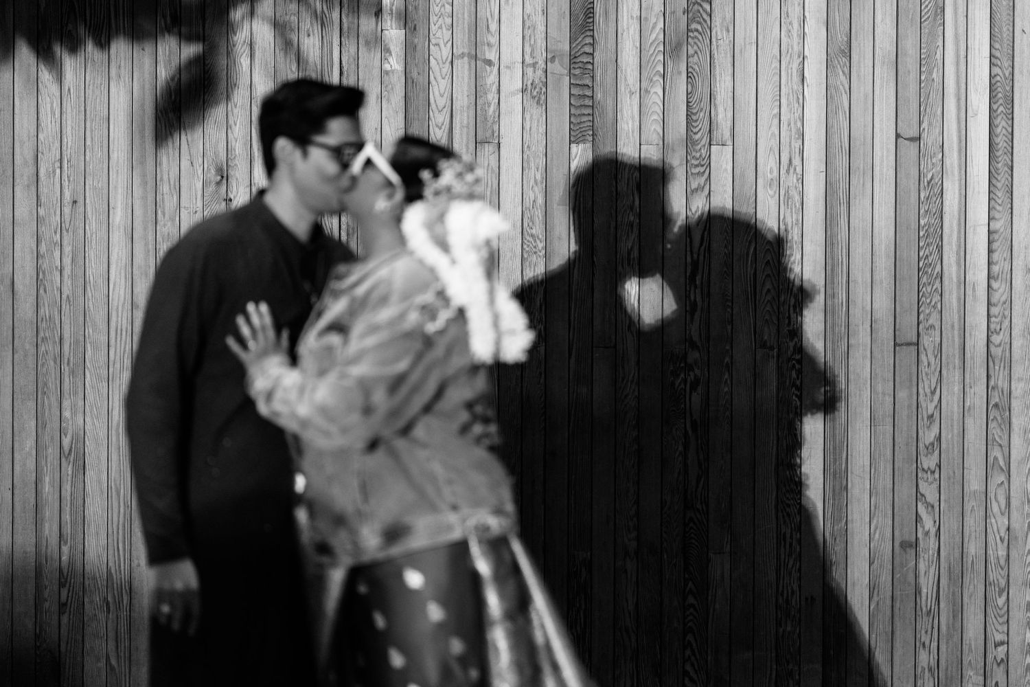 Black and white silhouette of couple kissing against wooden wall with dramatic shadows.