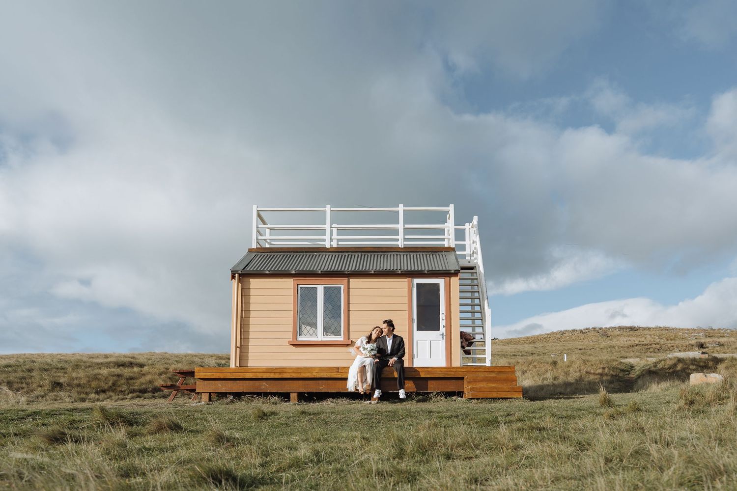 A small wooden cabin sits atop a grassy coastal hillside under blue skies.
