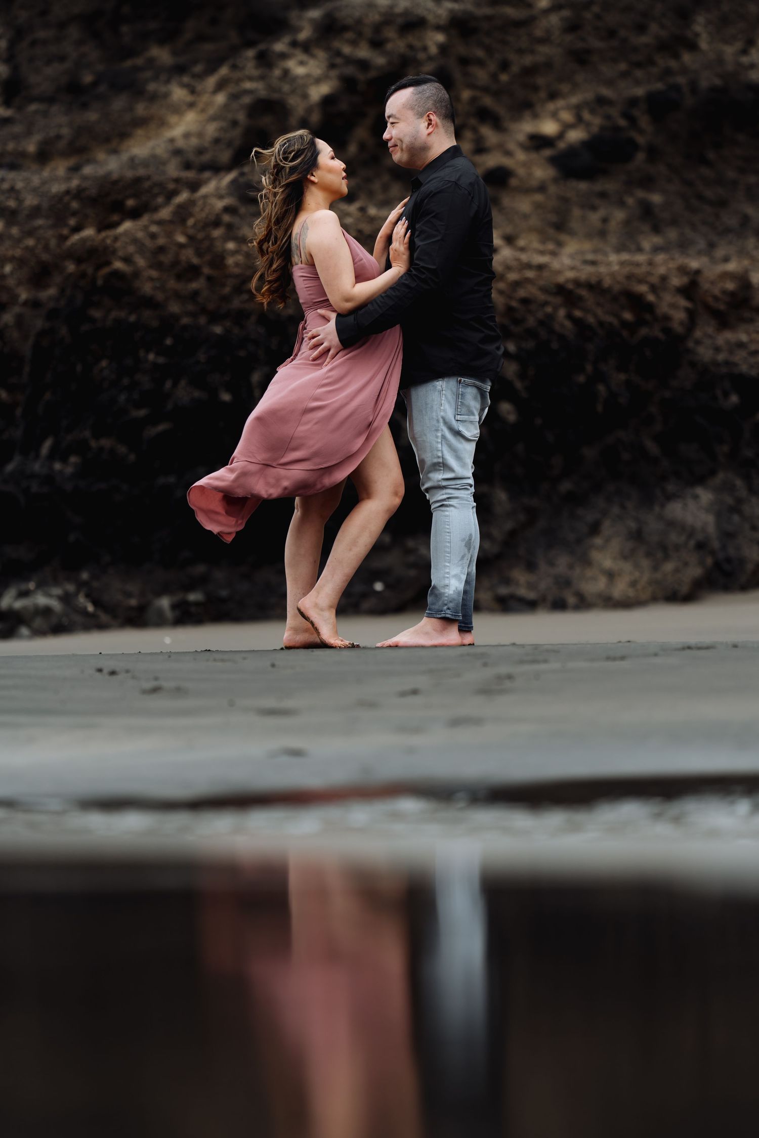 A couple dances together on a beach with flowing pink dress and dark cliffs behind them.