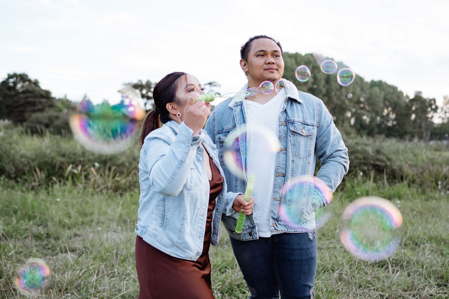 Playful couple blowing bubbles in outdoor field during sunset.