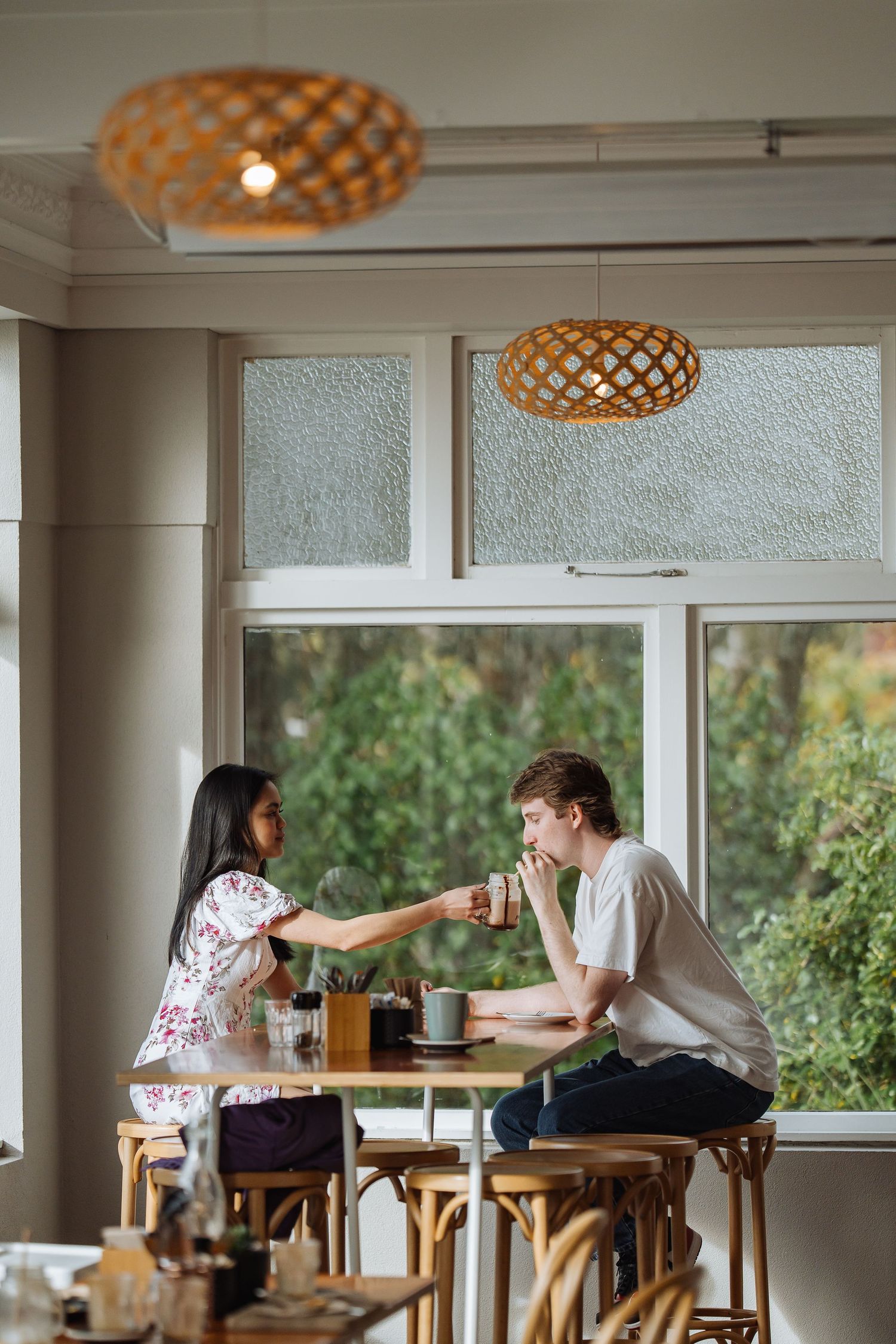Interior of bright cafe with wooden furniture and pendant lighting by large windows.