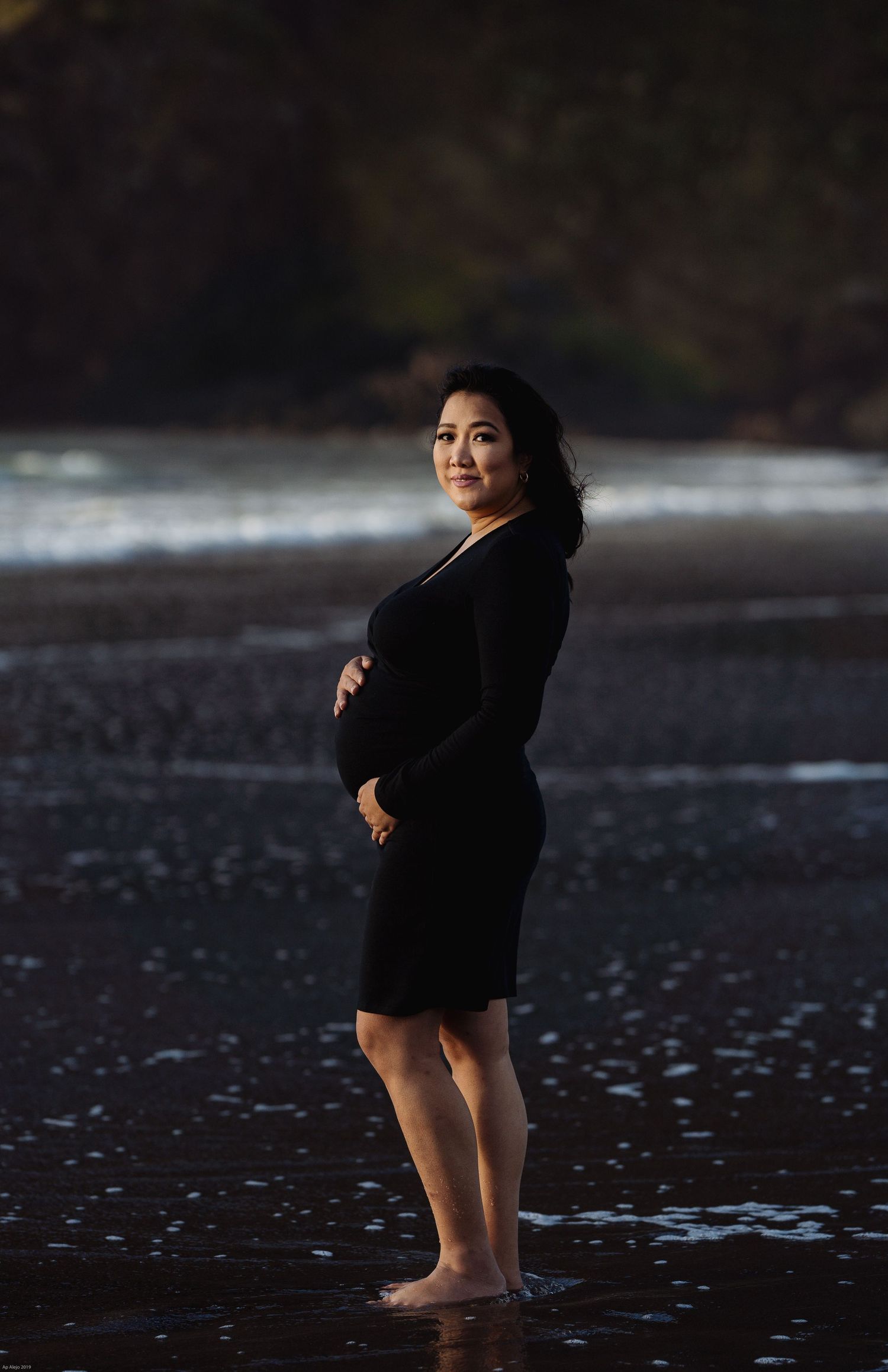 Figure in black dress stands barefoot on dark beach at sunset creating dramatic silhouette against ocean backdrop.