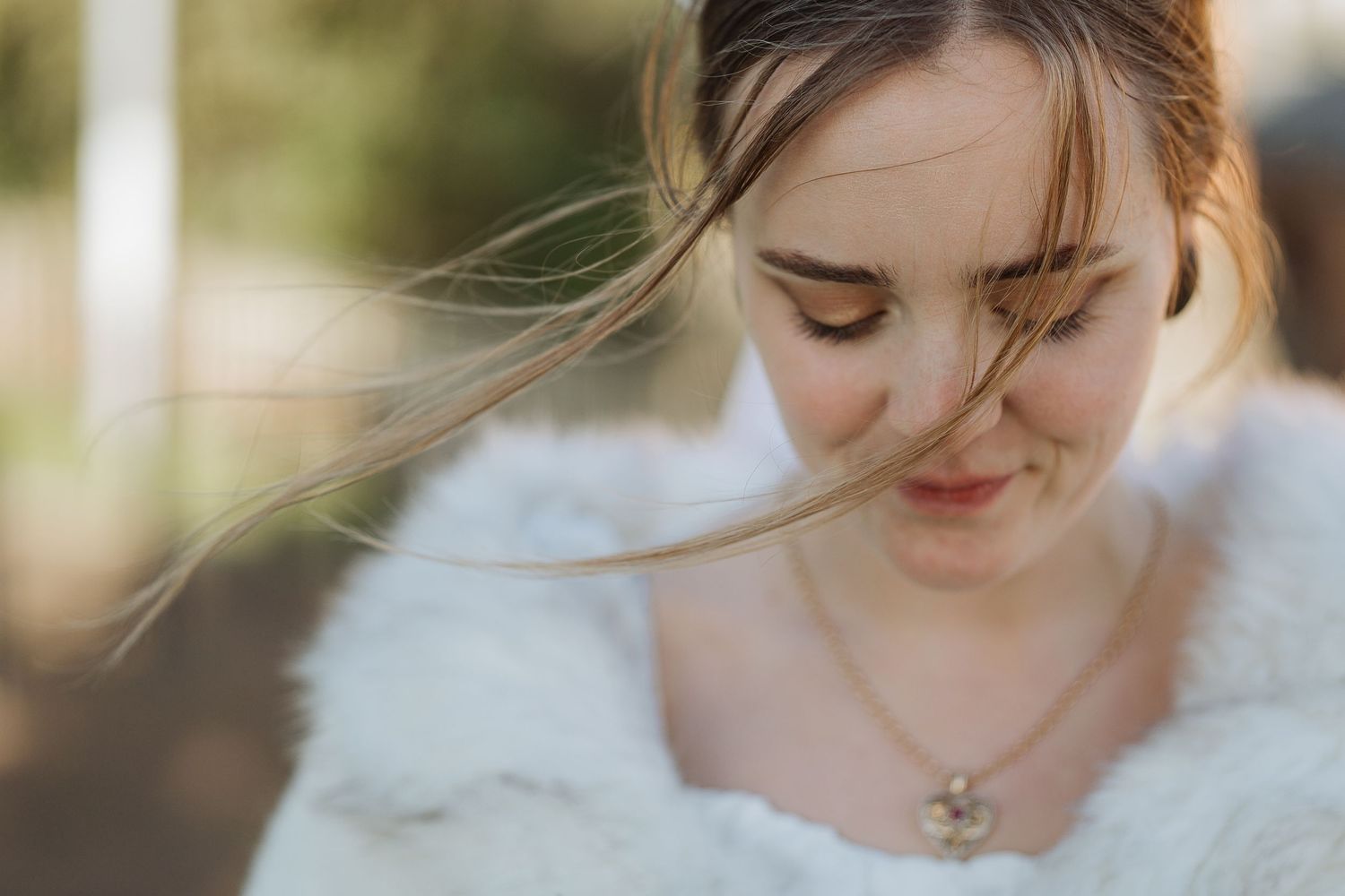 A bride in a white fur wrap smiles softly with windswept hair in warm sunlight.