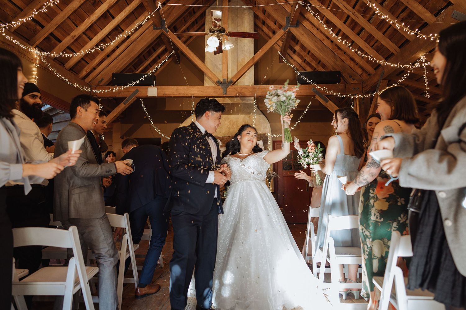 Wedding guests dance and celebrate under rustic wooden beams with string lights at a reception.