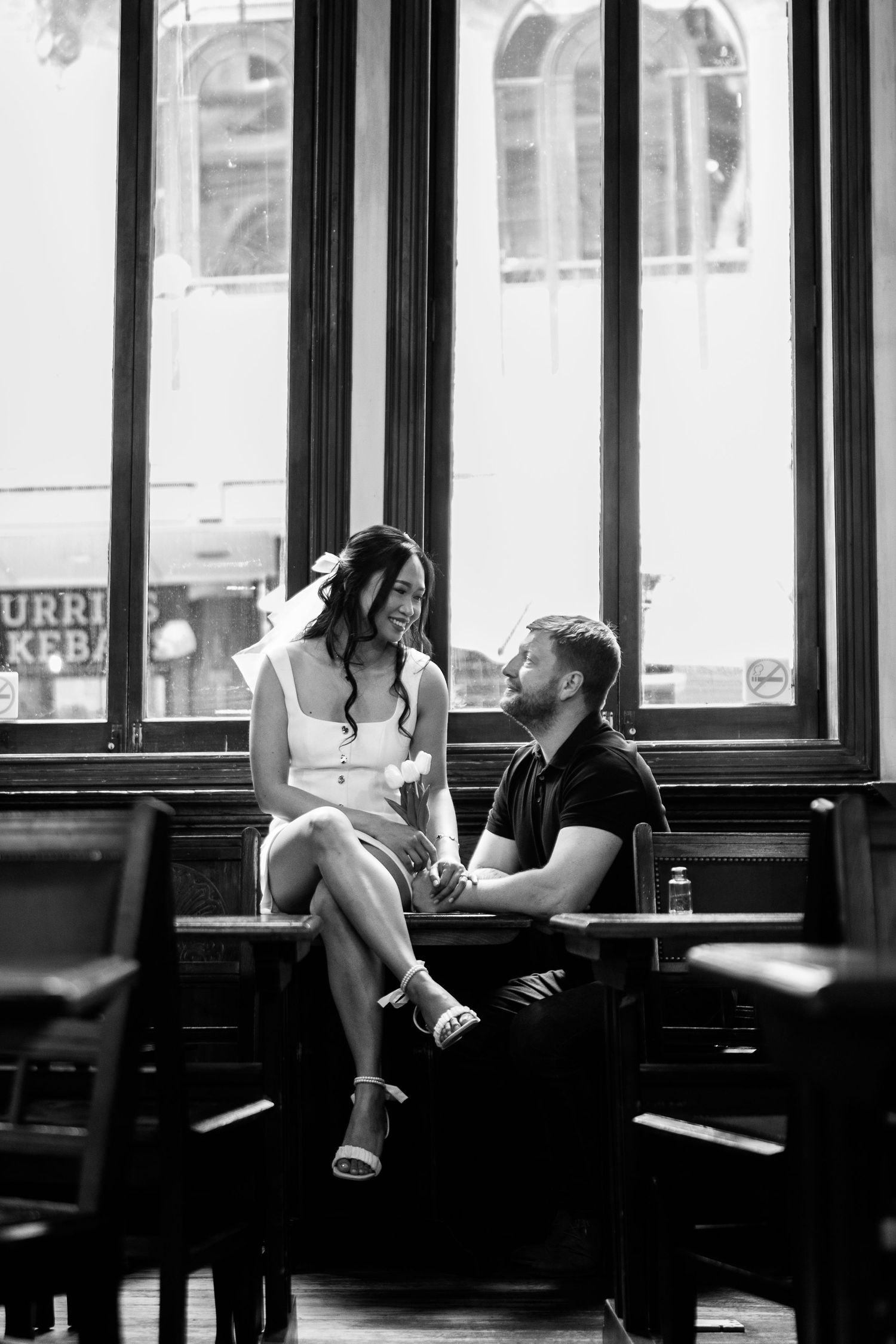 A couple sits together in silhouette on a dimly lit bench by large windows.