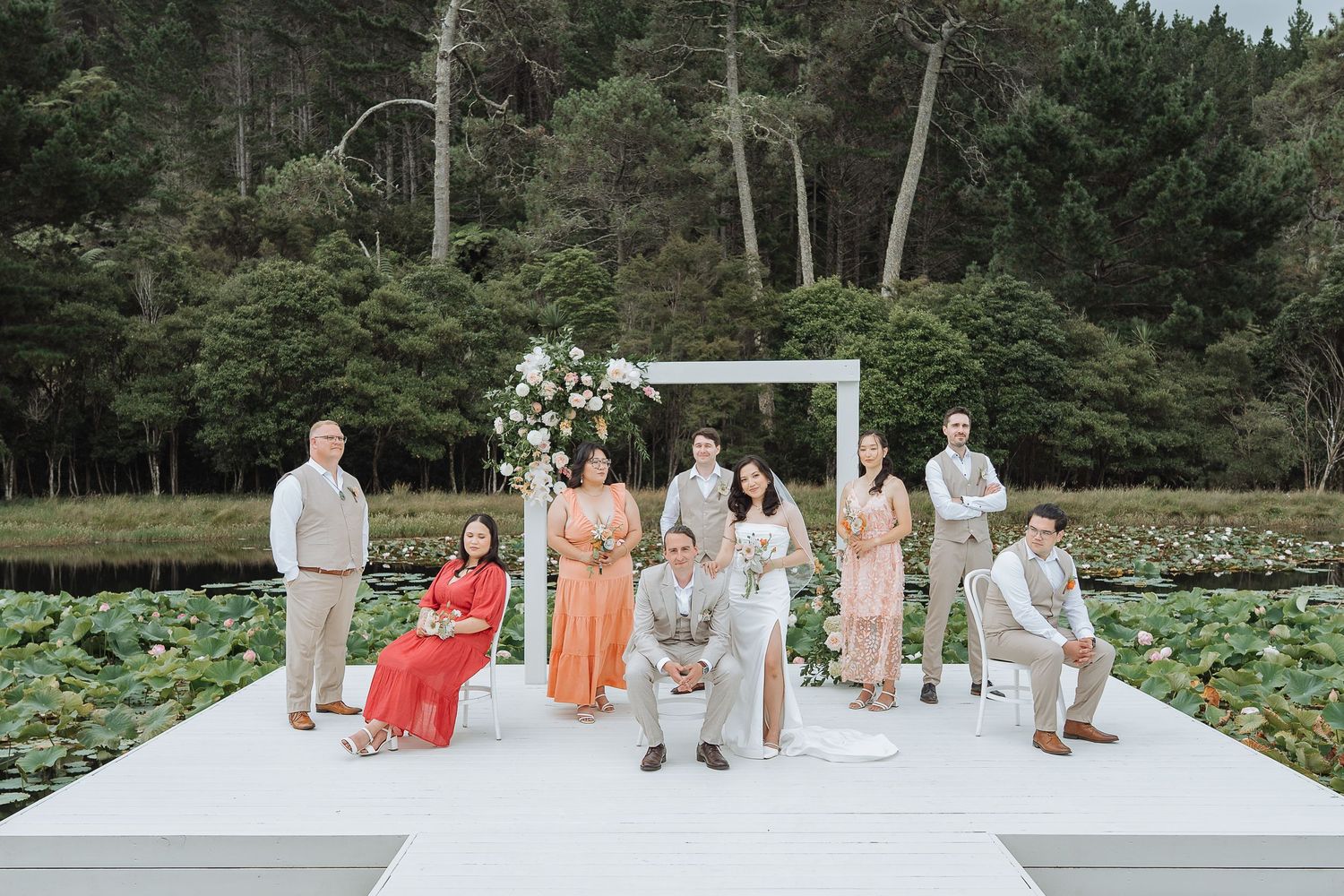 Wedding group poses on elevated white platform with forest backdrop and decorative floral arrangements.