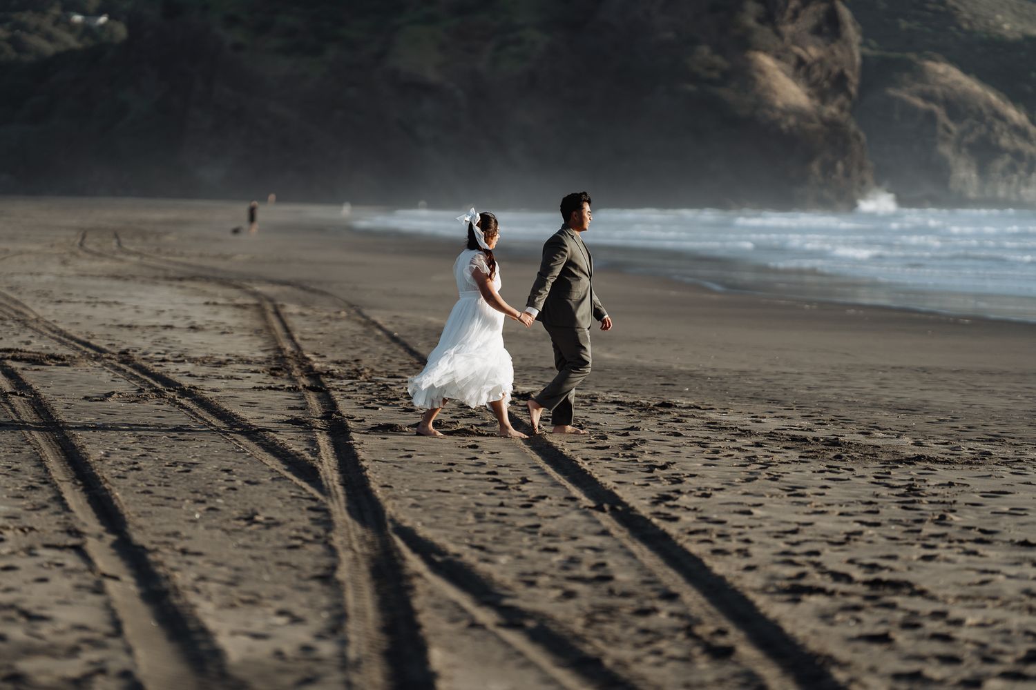 A couple walks along a scenic beach with mountains in the background during golden hour.