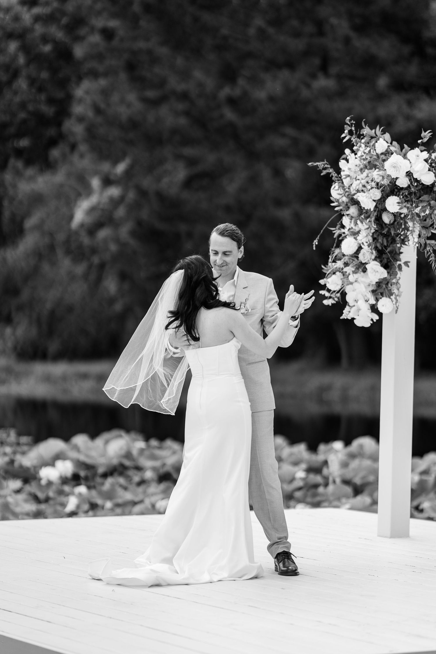 Black and white photo of a couple dancing during their wedding ceremony under a floral decorated arch.