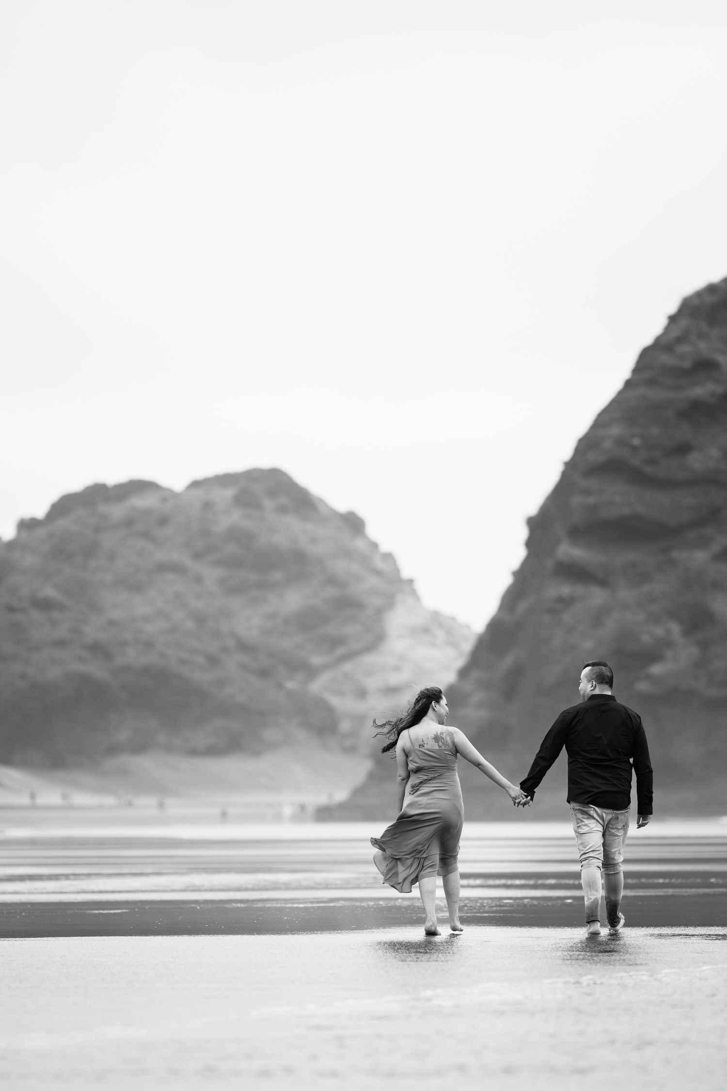 A black and white image of a couple walking together on a beach with towering cliffs.