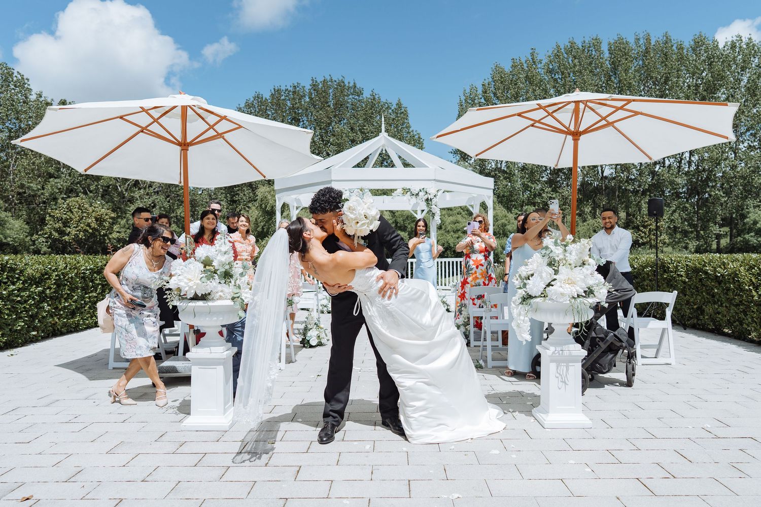 Outdoor wedding reception setup with white umbrellas and tables on sunny patio.