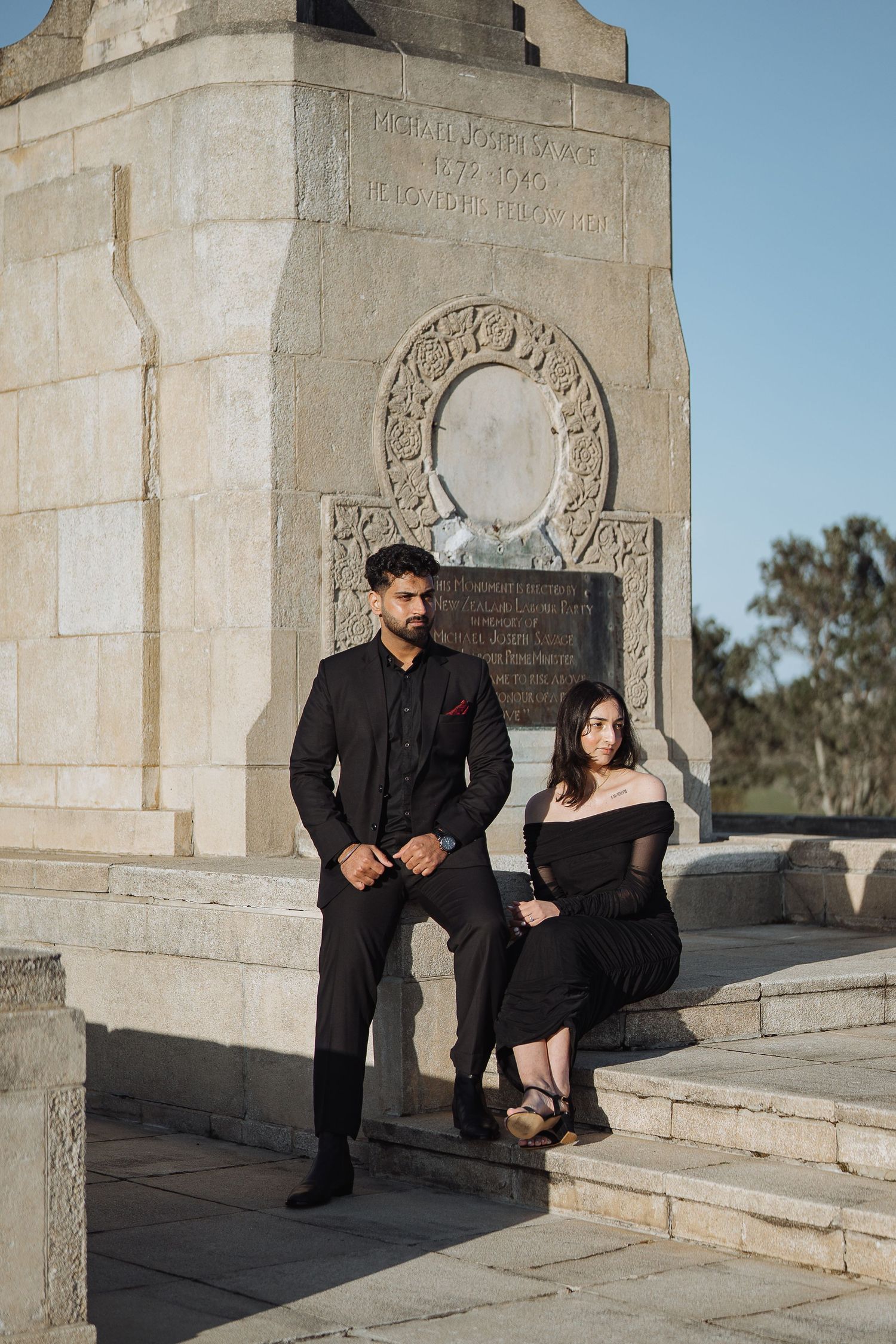 A couple poses against stone monuments in formal black attire.