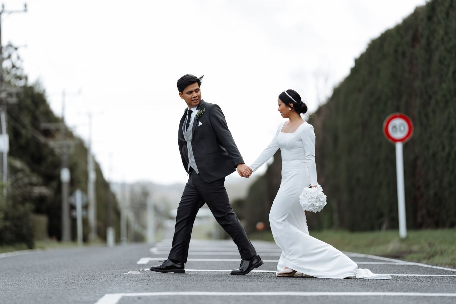 A couple in wedding attire poses playfully on a street with tall hedges and road signs.
