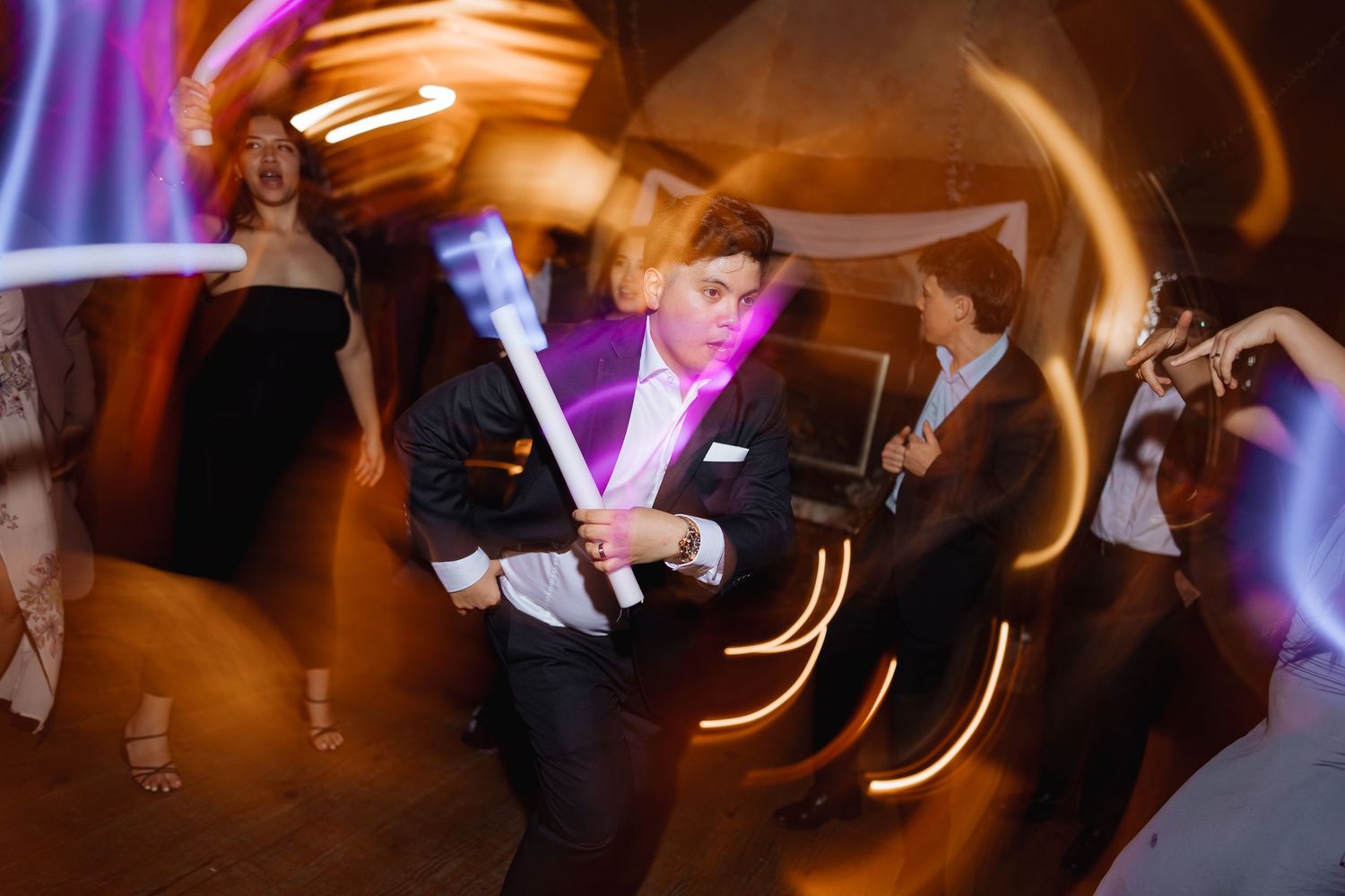 Motion-blurred dancers create light trails under purple spotlights at a lively wedding reception.