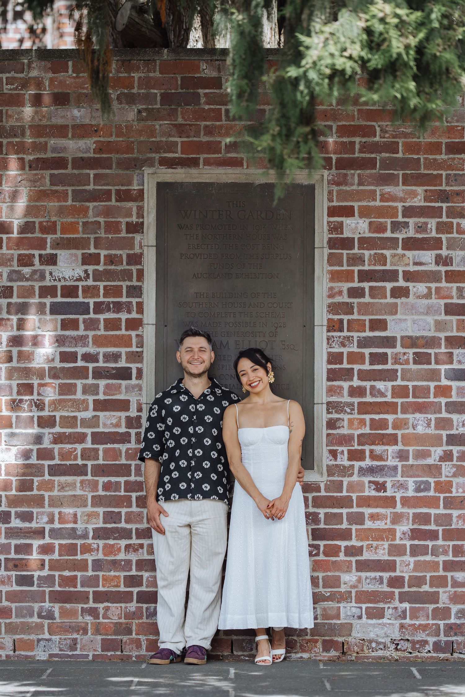 A couple stands together against a brick wall backdrop under hanging foliage.