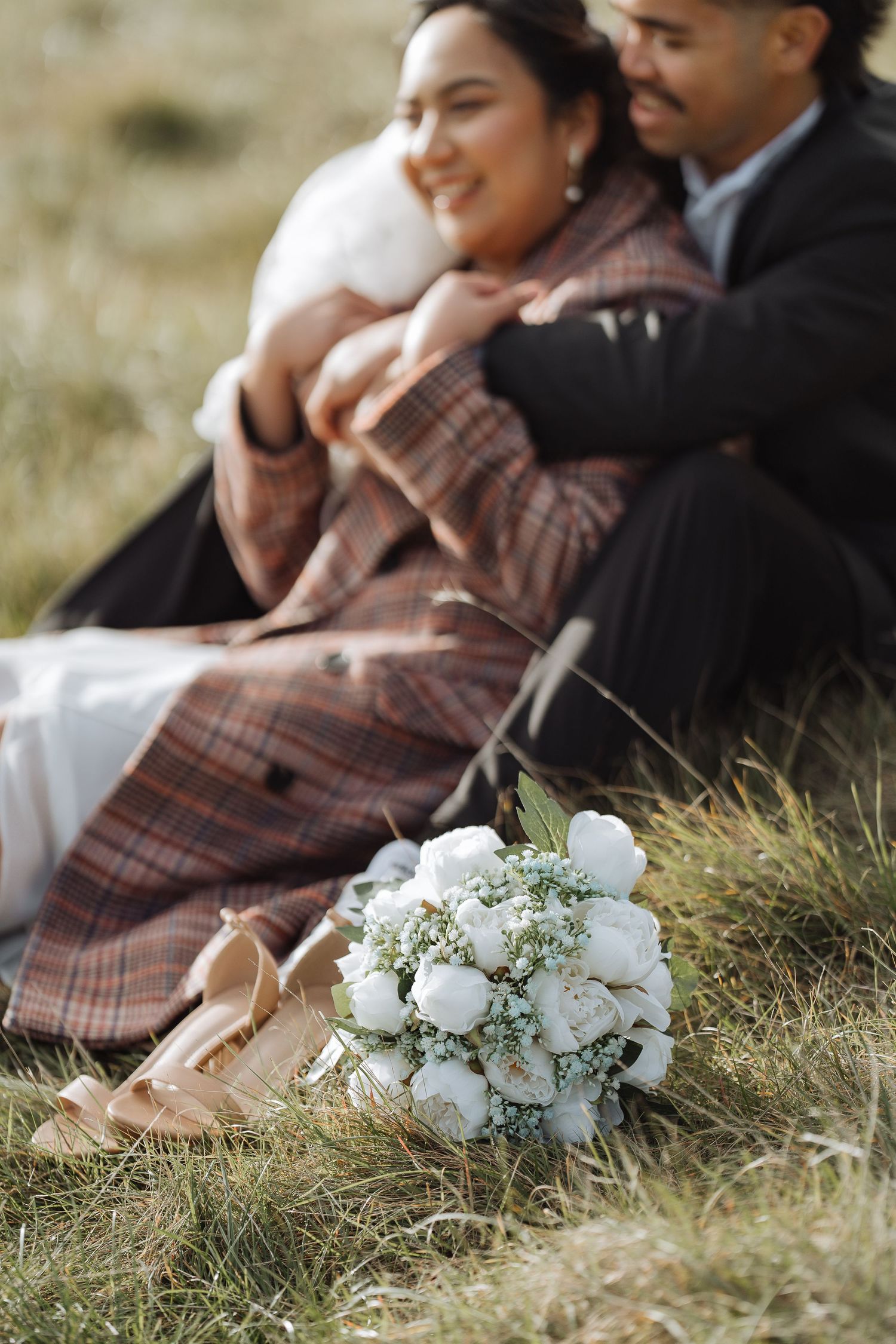 A white floral wedding bouquet rests on grass next to a plaid blanket.