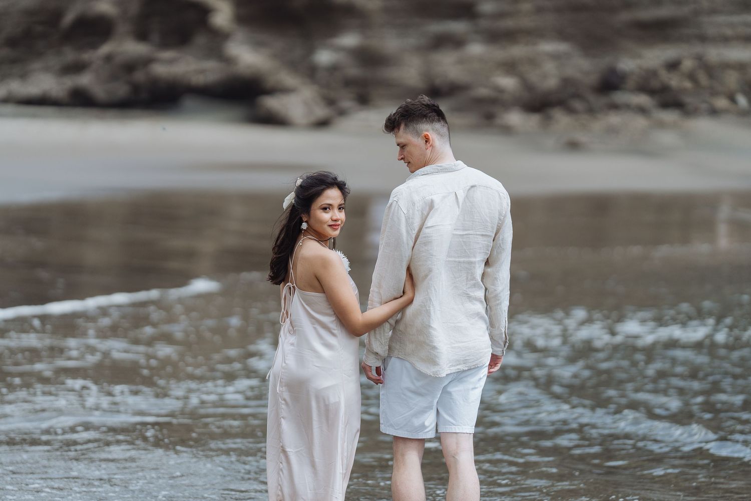 Intimate beach portrait session with couple in white attire against rocky shoreline.