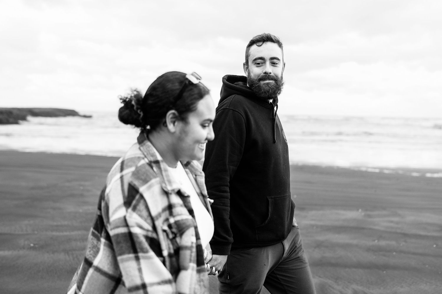 Black and white photo of a bearded man and woman walking hand in hand on a beach with ocean waves behind them.