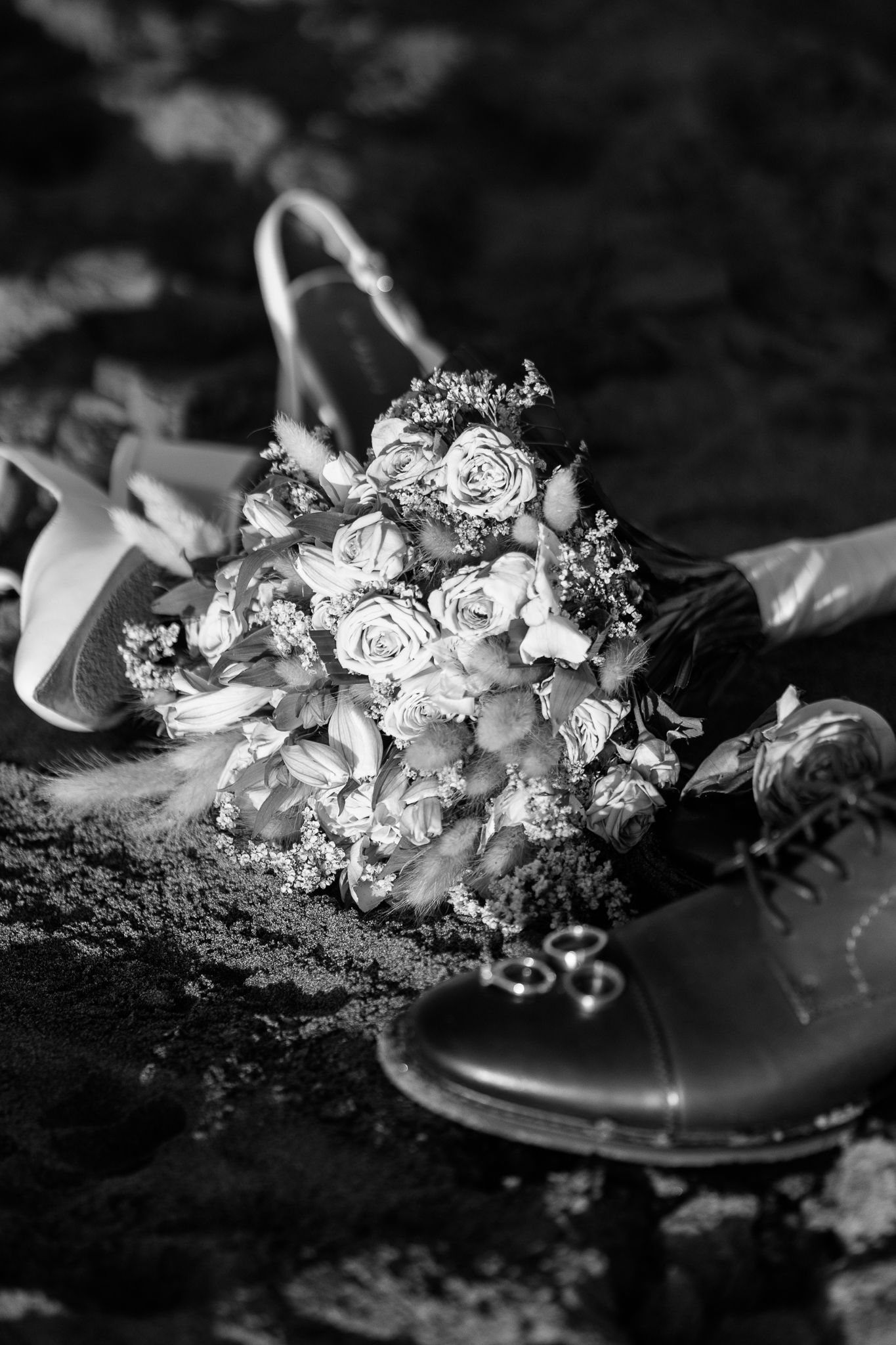 Black and white wedding detail photo of bridal bouquet and shoes on textured surface.