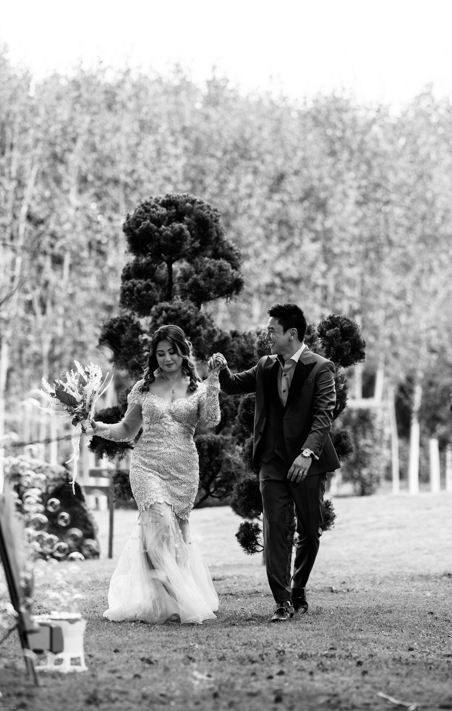 Black and white photo of newlyweds walking together in garden with decorative black pom poms.