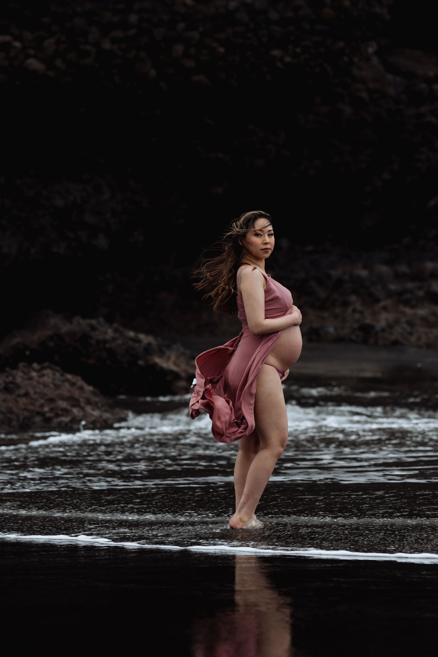A maternity photo of a person in a flowing pink dress standing at the edge of dark waters.